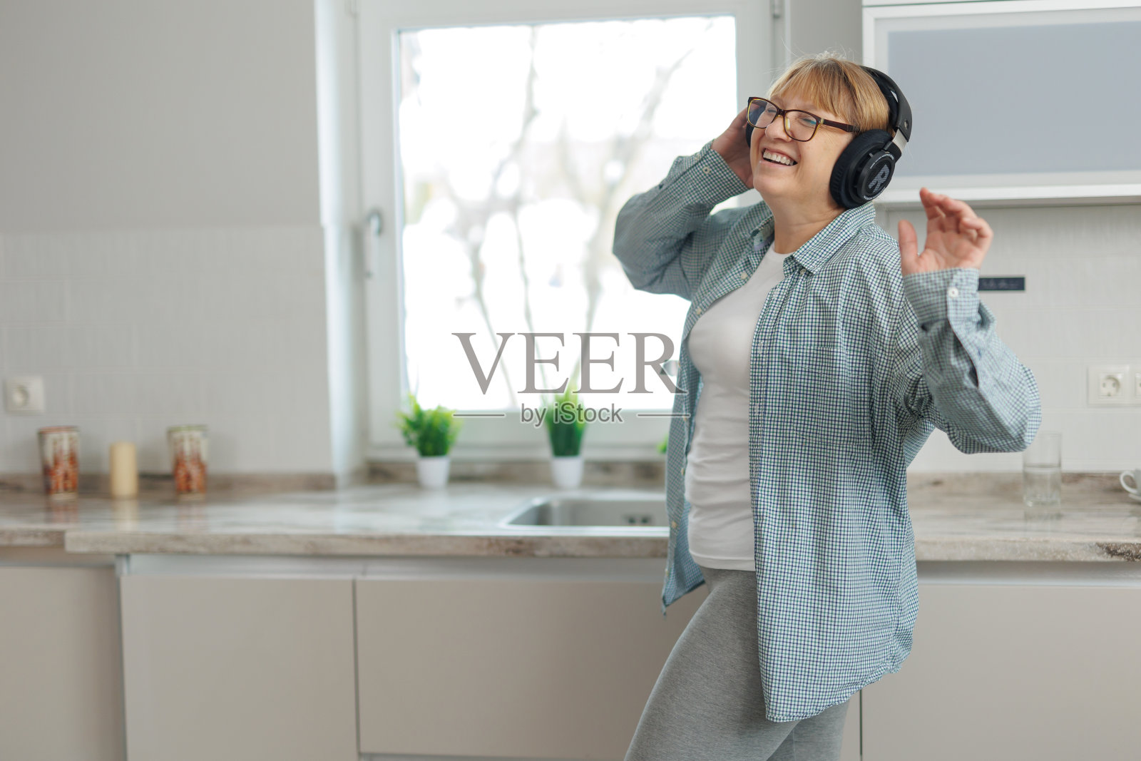 一名成年女子带着无线耳机在厨房里听音乐跳舞。一个穿着蓝色衬衫的女人微笑着跳舞。照片摄影图片