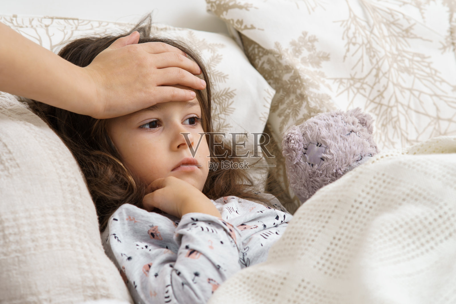 不认识的女人在卧室摸小女孩的额头。无聊的女孩和熊玩具躺在白色柔软的床上照片摄影图片