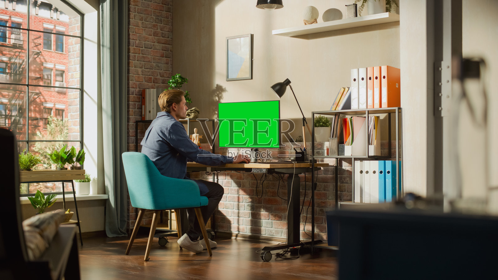 年轻英俊的男子从家里工作在台式电脑与绿色屏幕模拟显示。检查社交媒体，浏览互联网。明亮阁楼公寓的客厅。照片摄影图片