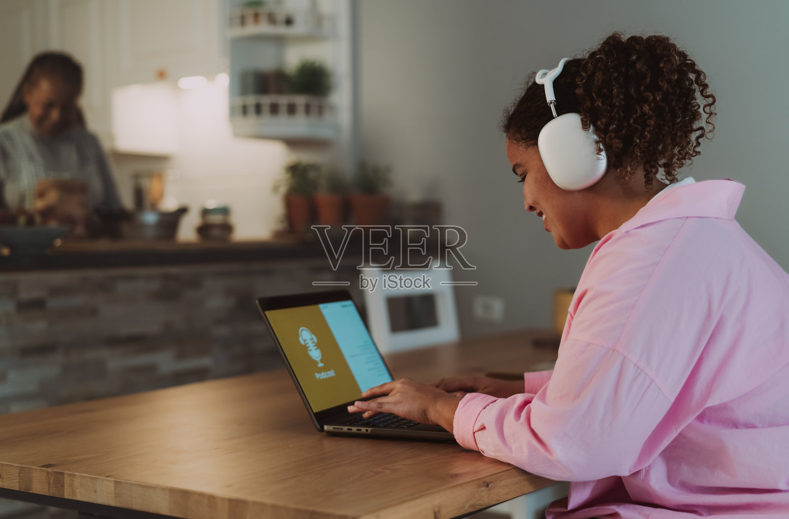 年轻的非洲女孩使用笔记本电脑听播客，而她的母亲在家里做饭的背景照片摄影图片