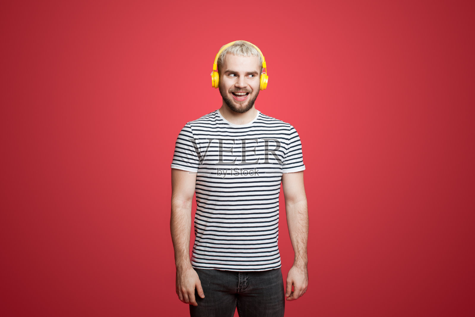 肖像逗趣和快乐的年轻人听音乐耳机，微笑高兴，站在红色背景。享受他最喜欢的音乐。照片摄影图片