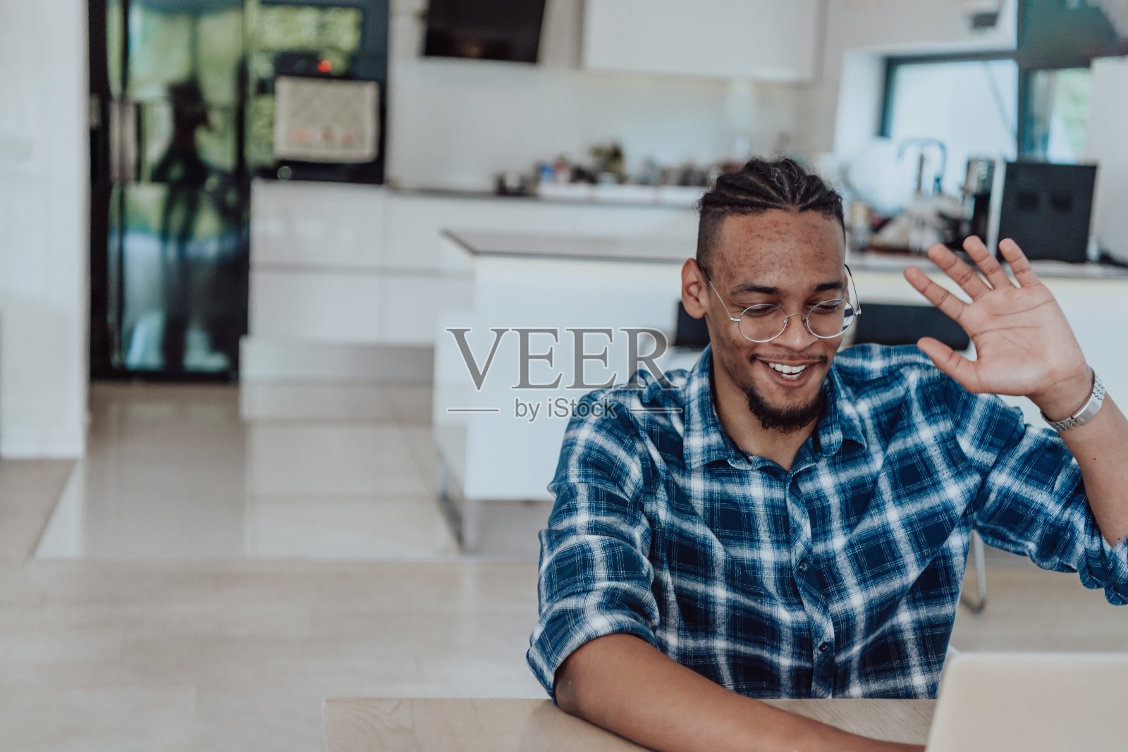 一名戴眼镜的非裔美国男子坐在现代客厅的一张桌子旁，用笔记本电脑进行商务视频聊天，与朋友交谈和娱乐照片摄影图片