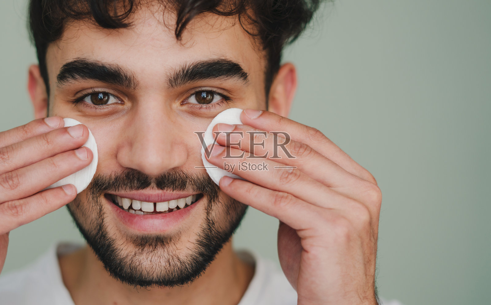 特写肖像的一个有吸引力的微笑男子手持化妆棉在他的脸颊上应用乳液孤立在粉彩背景工作室。皮肤护理保健美容程序概念照片摄影图片