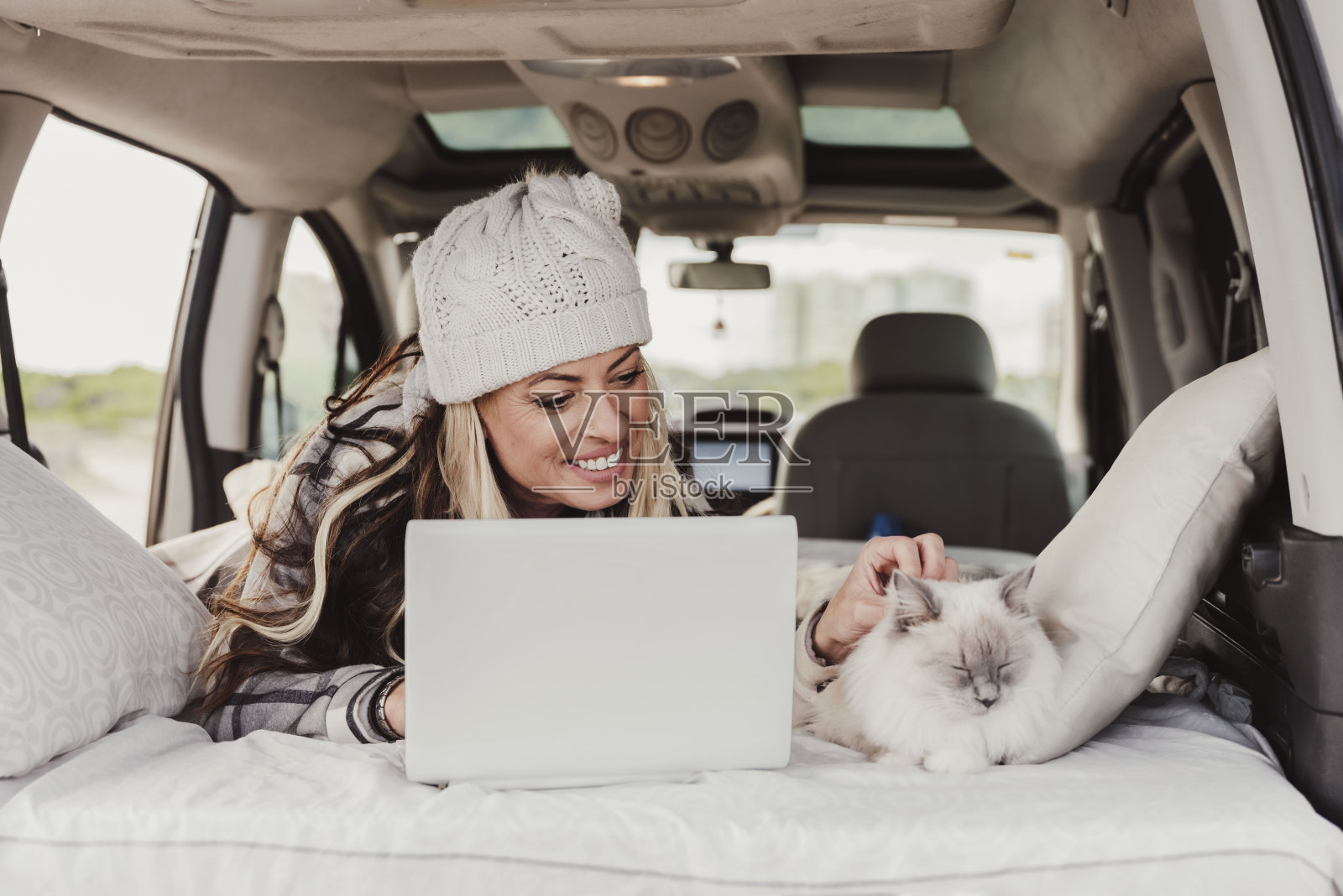 带着绒线帽的女人和猫躺在露营车里用笔记本电脑。数字游牧生活方式照片摄影图片