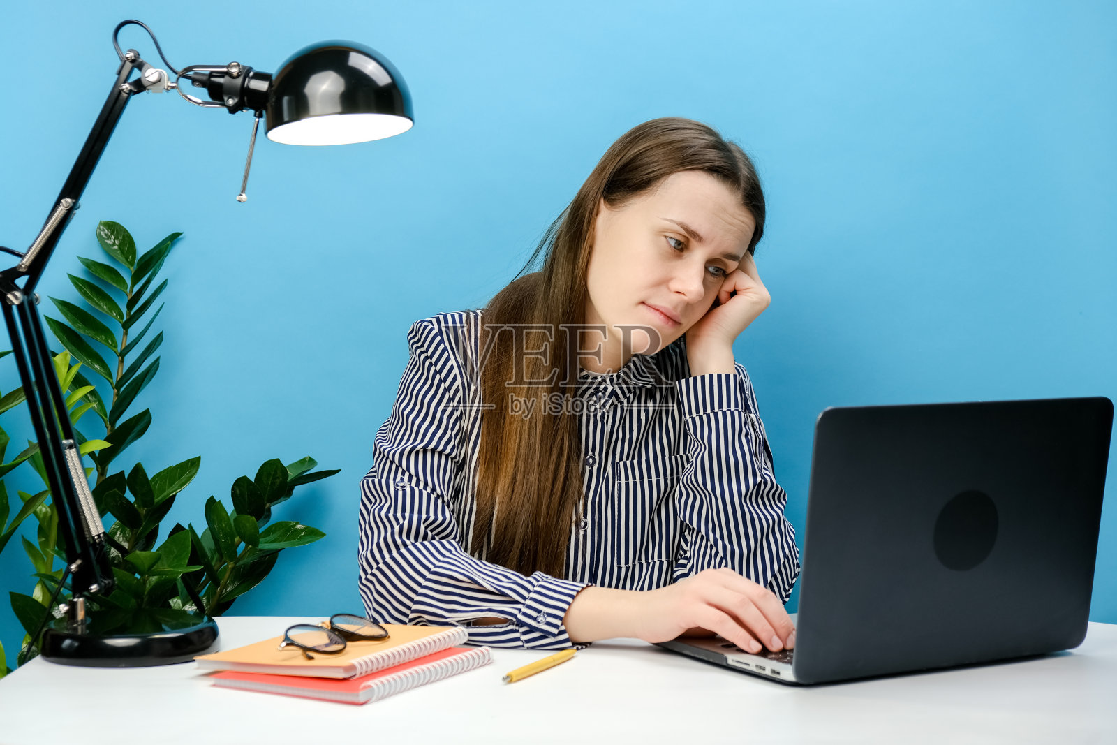 困惑疲惫沮丧的秘书员工年轻女性20多岁，穿着衬衫坐在白色办公桌前工作，使用笔记本电脑上网支撑脸，在工作室的蓝色背景墙上摆出孤立的姿势照片摄影图片