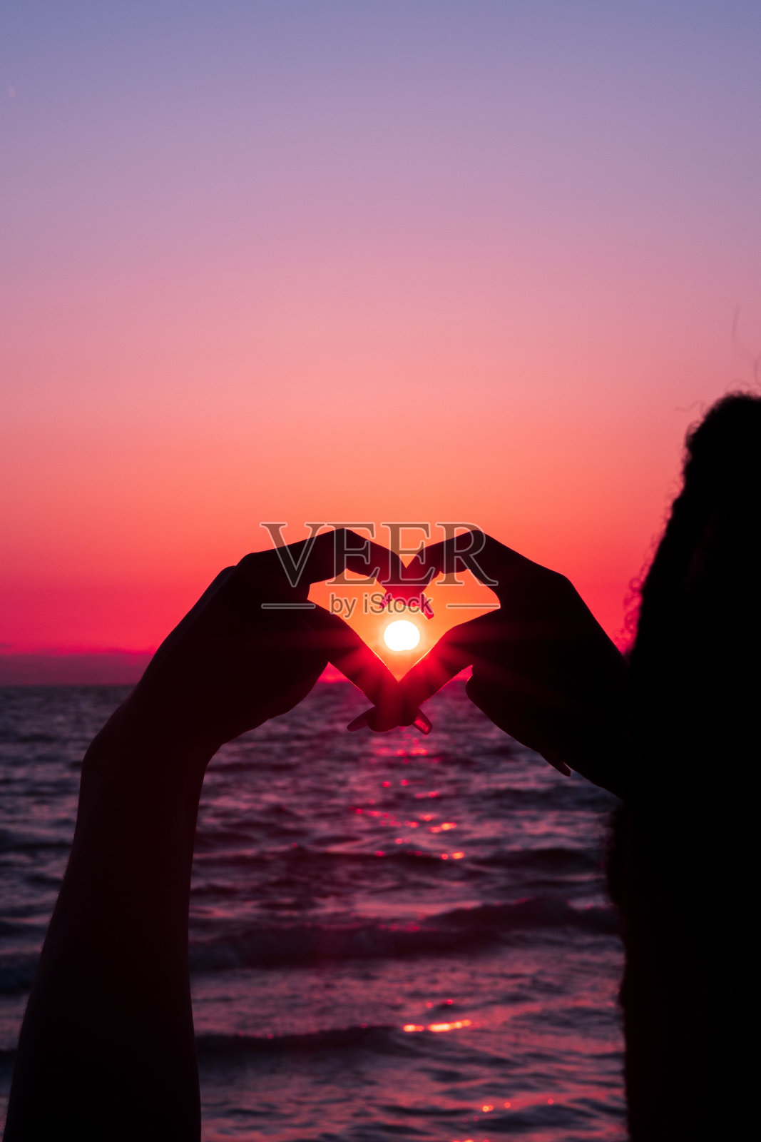 一个女孩的剪影在日落的海滩上，用手指围绕着太阳做心脏照片摄影图片