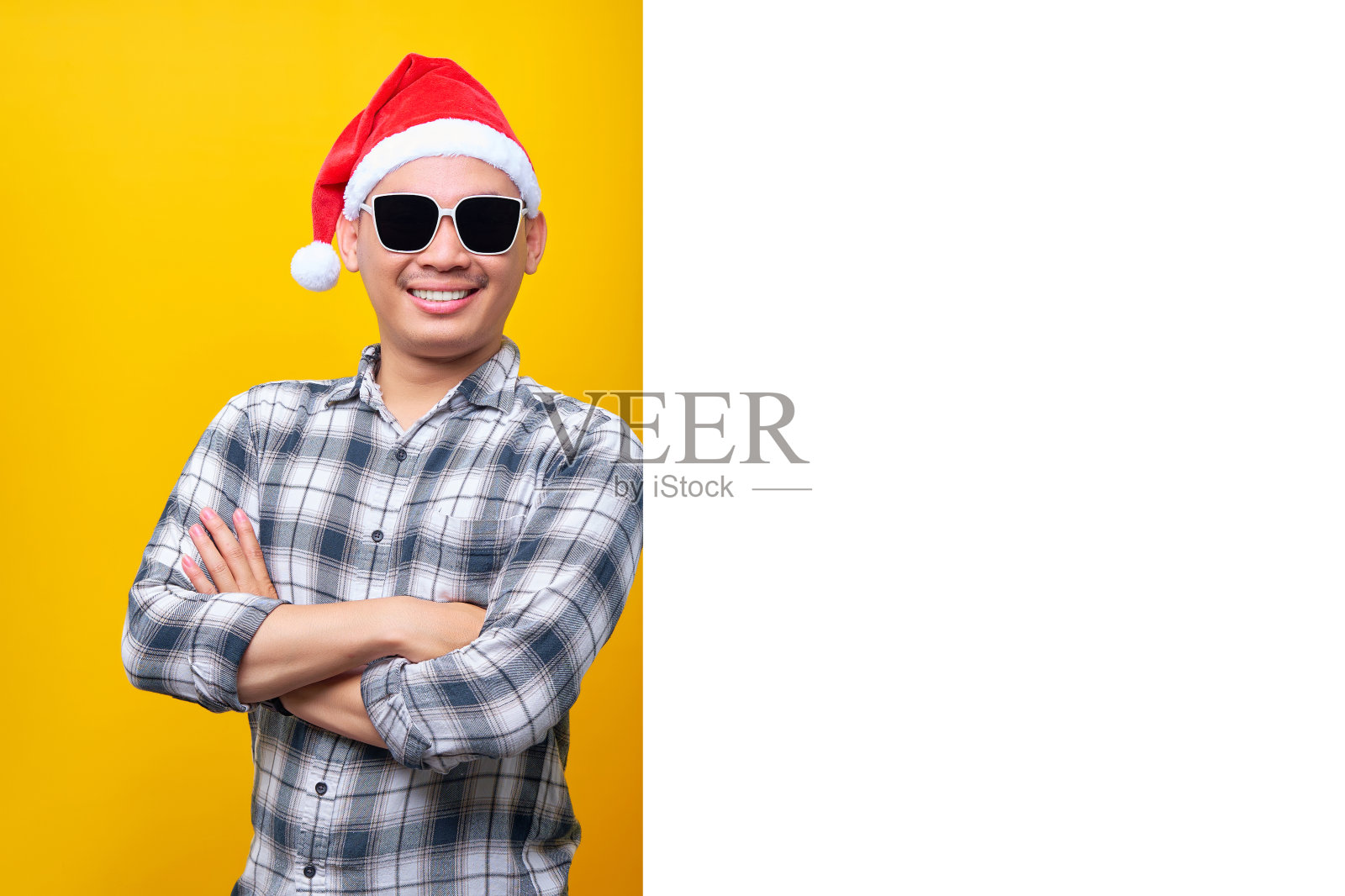 开朗的年轻亚裔男子戴着圣诞帽和眼镜，站在黄色背景上孤立的一面白色广告牌上。2023年新年快乐假期概念照片摄影图片
