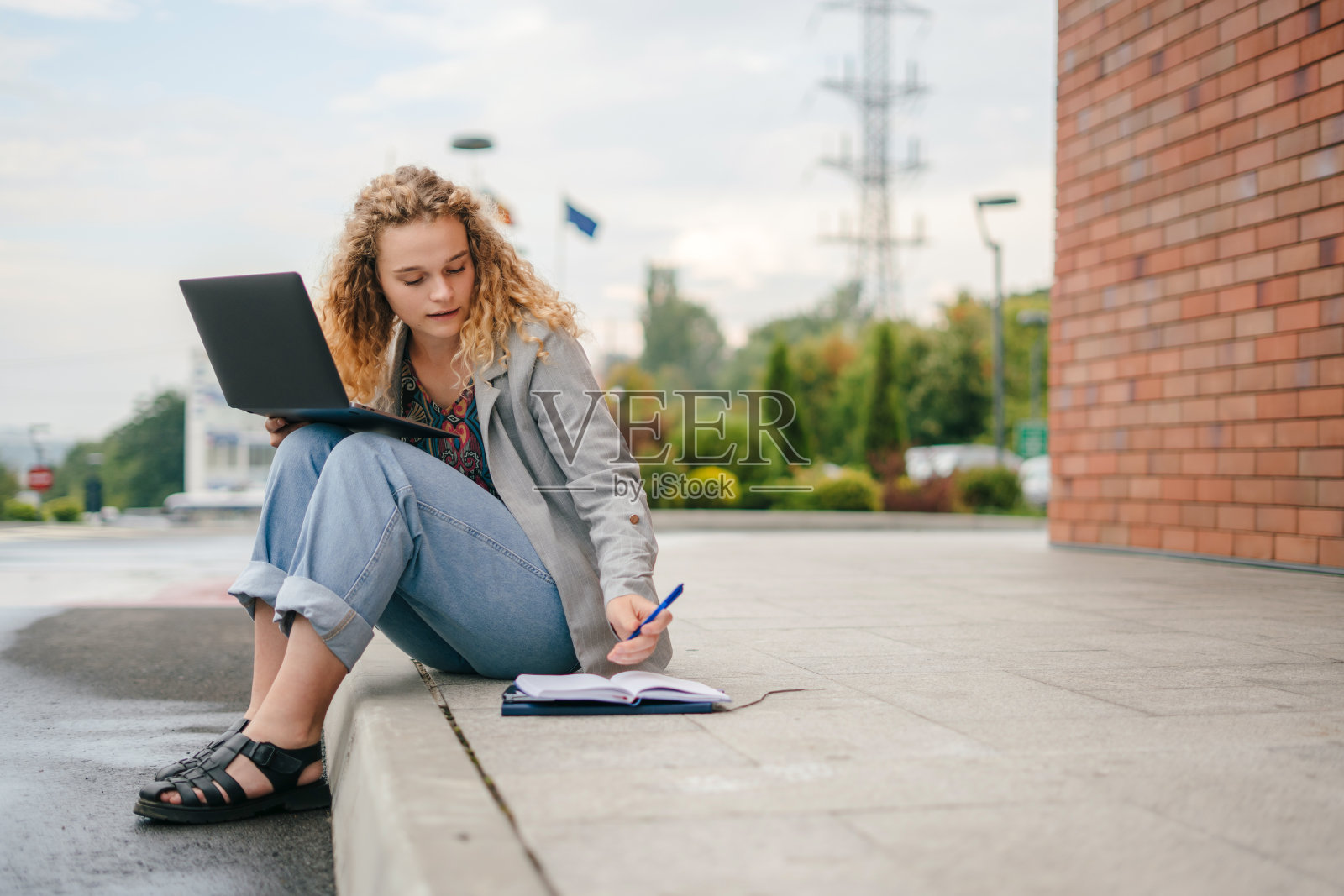 快乐的千禧一代女孩坐在街上用笔记本电脑做大学项目，在她的笔记本上写笔记。学生在线学习，工作。照片摄影图片