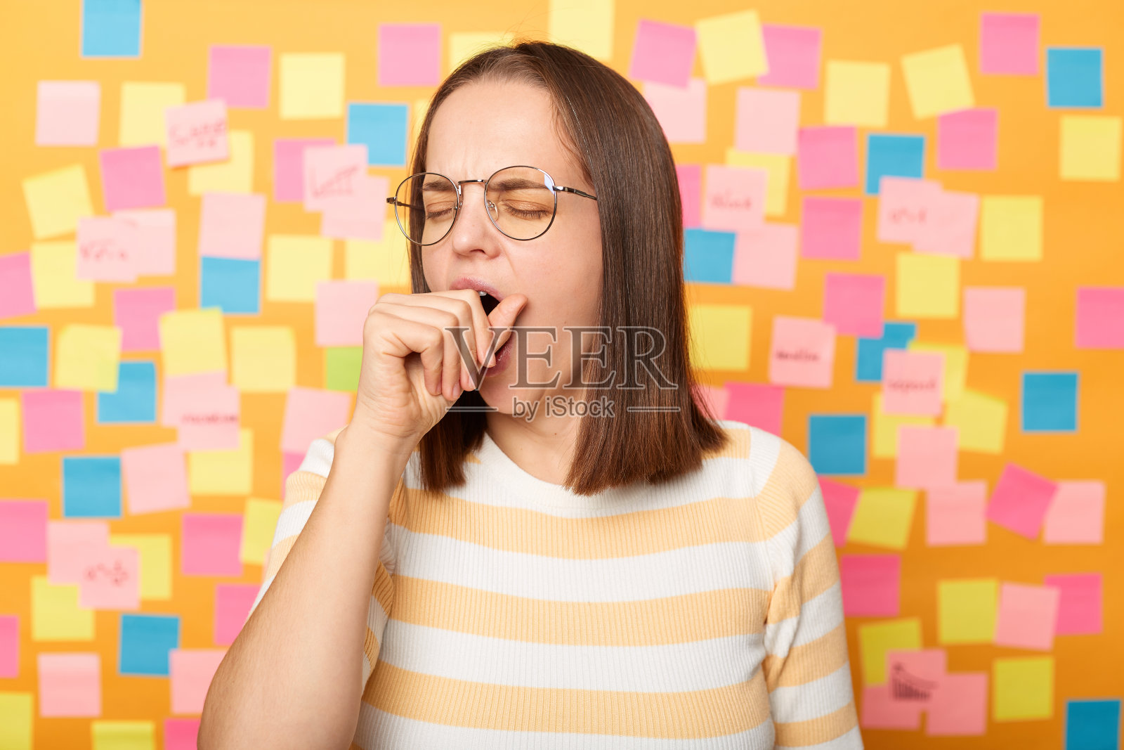 肖像嗜睡过度工作的有吸引力的女人穿着条纹t恤站在黄色墙壁覆盖的贴纸，打哈欠，用拳头覆盖嘴，保持闭着眼睛。照片摄影图片