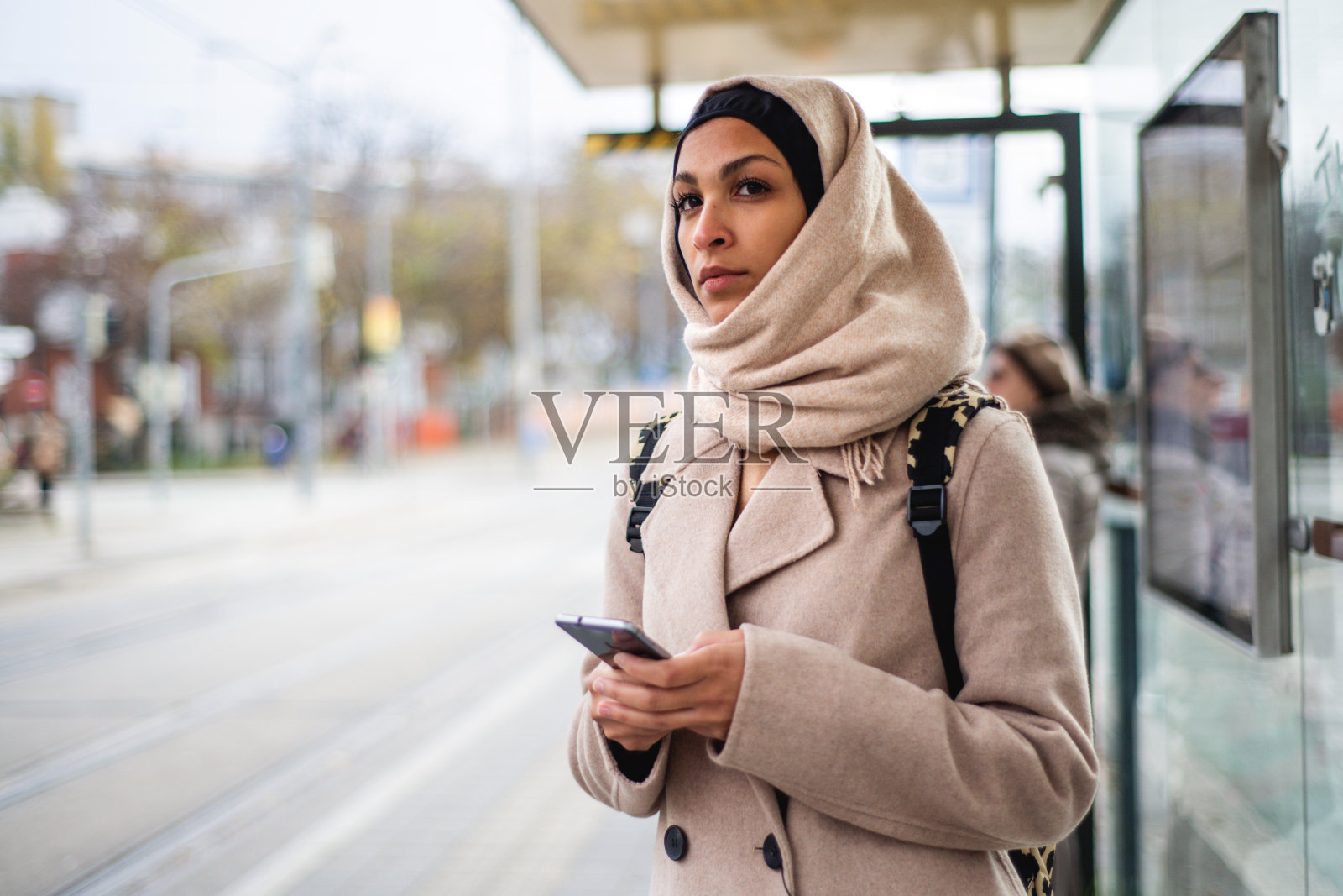 年轻的穆斯林妇女拿着智能手机在城市公交车站等车。照片摄影图片