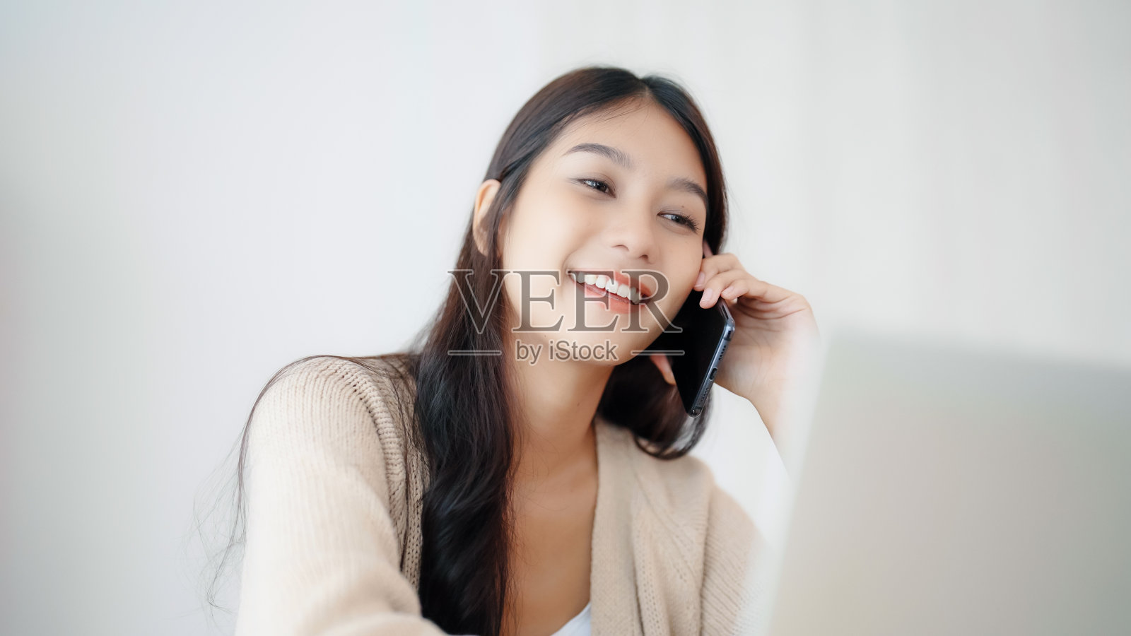 年轻的亚洲女子一边在家里客厅的桌子上用笔记本电脑工作，一边说着智能手机照片摄影图片