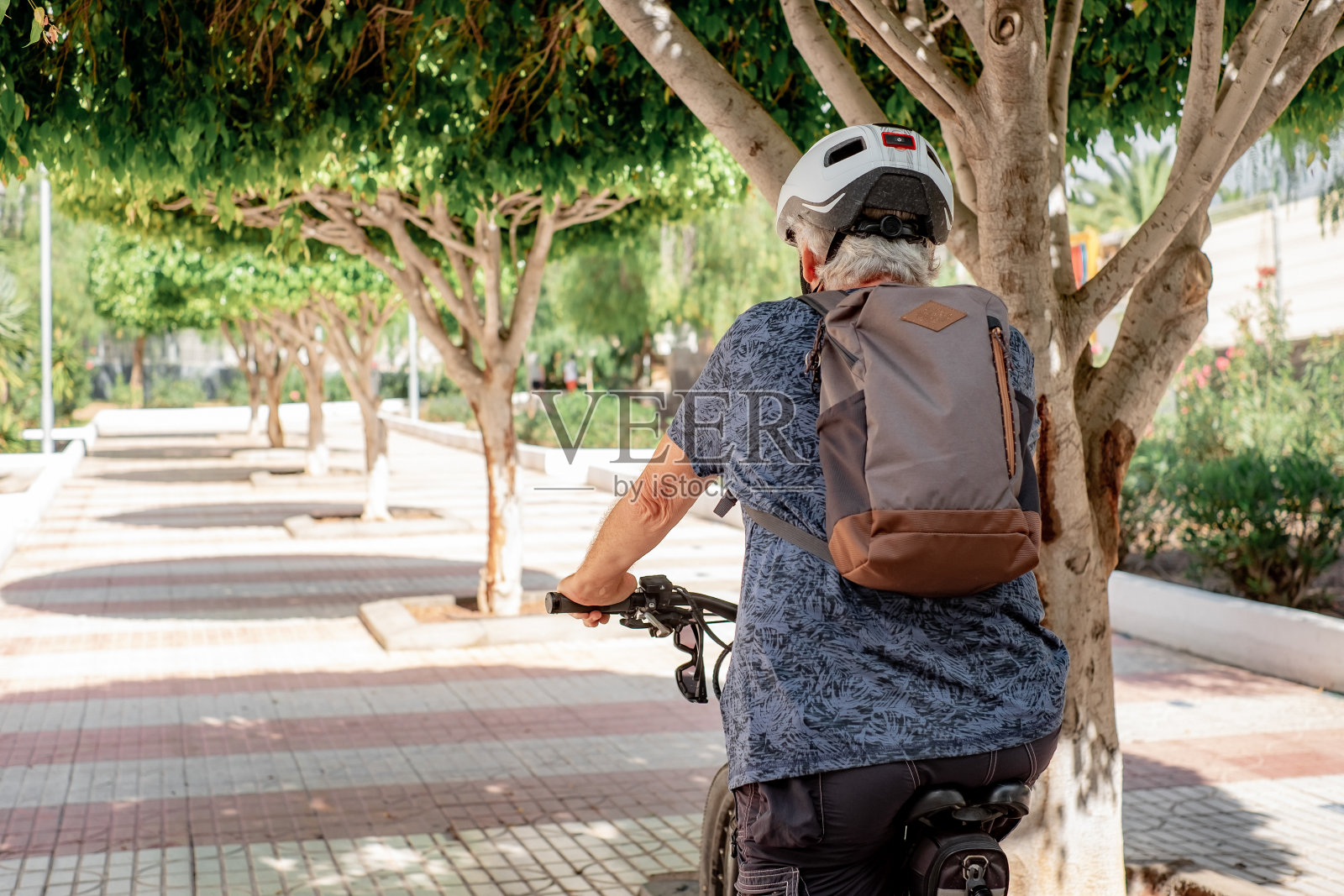 背着头盔和背包的老年活跃男子在城市公园用电动自行车跑步的背影。白人老人享受健康的生活方式和退休照片摄影图片