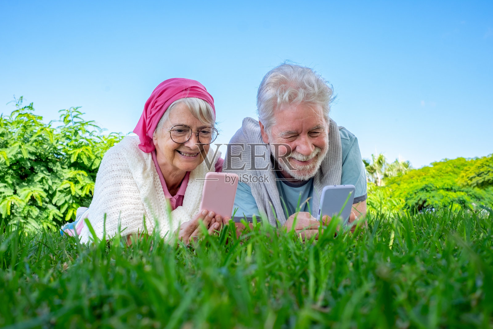 老人微笑着躺在公园的草地上，看着他们的手机。浪漫的现代夫妇享受自由时间和退休照片摄影图片