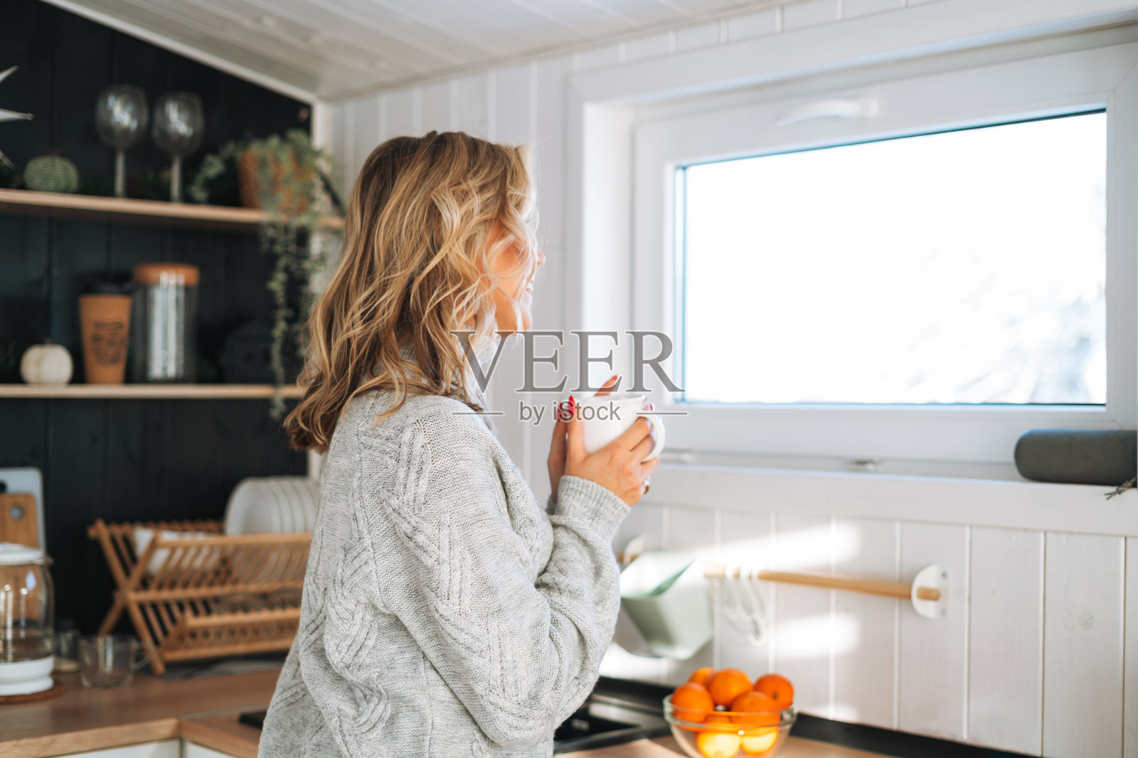 身穿灰色毛衣的年轻女子手里拿着一杯热饮，看着房子里厨房的窗户照片摄影图片