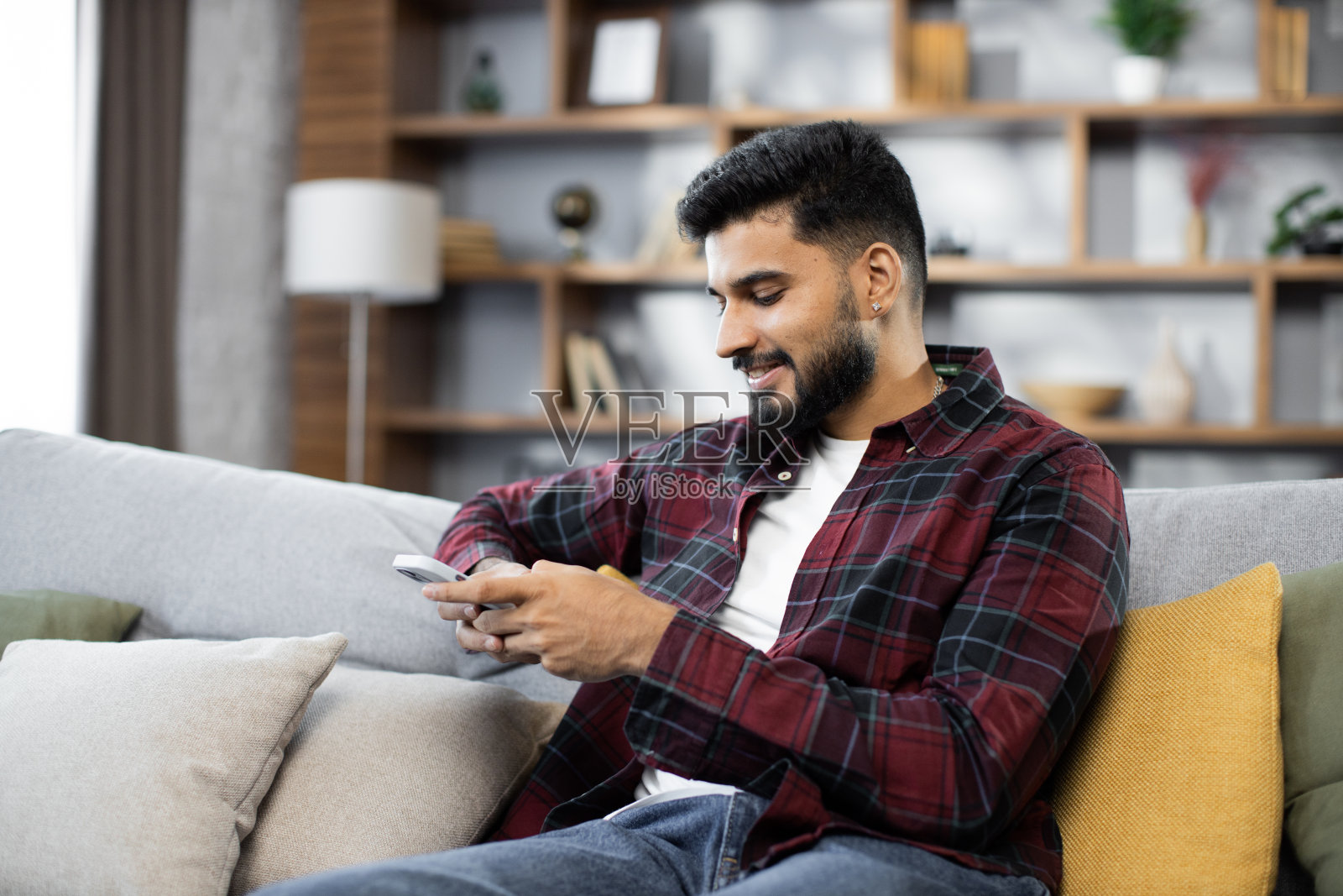 穿着红色格子衬衫的迷人男子坐在沙发上，在室内使用现代智能手机。照片摄影图片