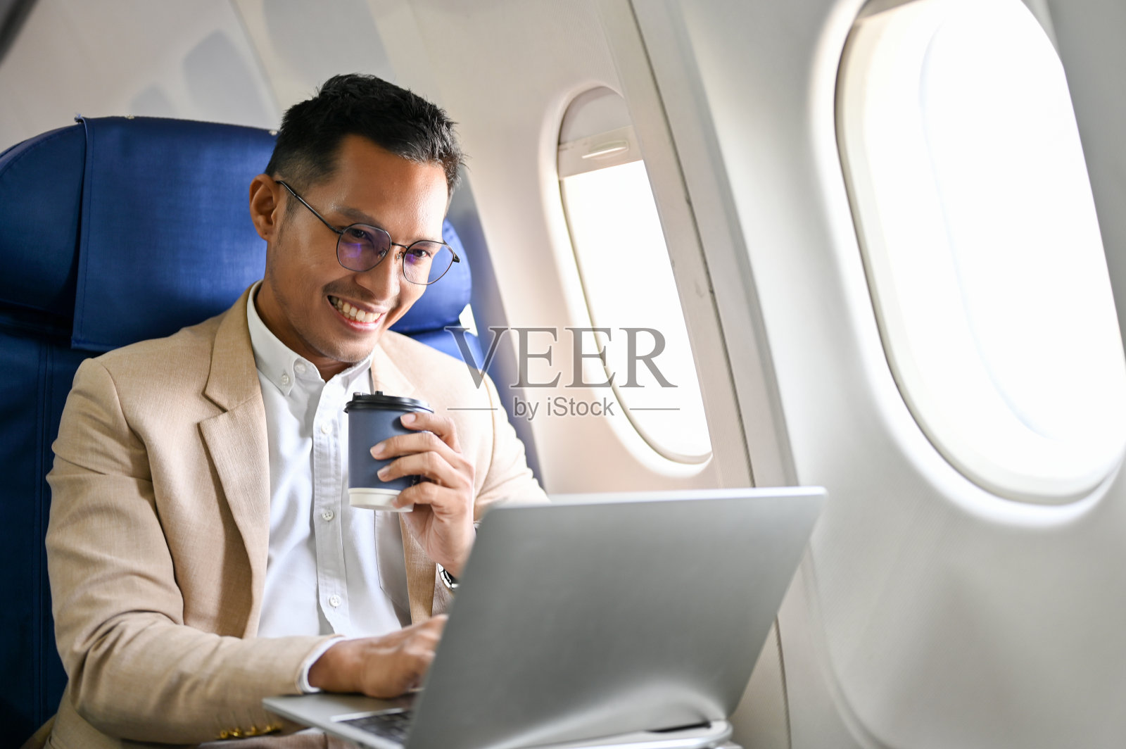 精明的亚洲商人一边喝着咖啡，一边用笔记本电脑管理飞行中的任务。照片摄影图片