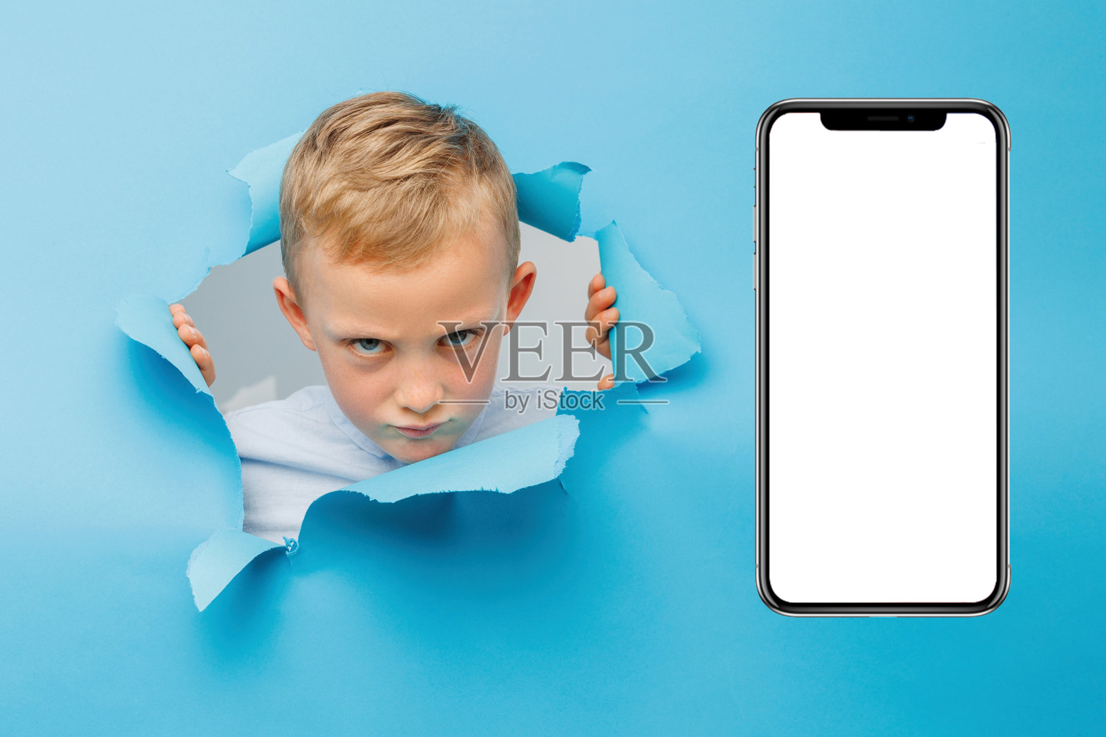 愤怒的可爱男孩在蓝色背景墙上玩得很开心，爬过一个巨大的手机旁边的纸洞。照片摄影图片