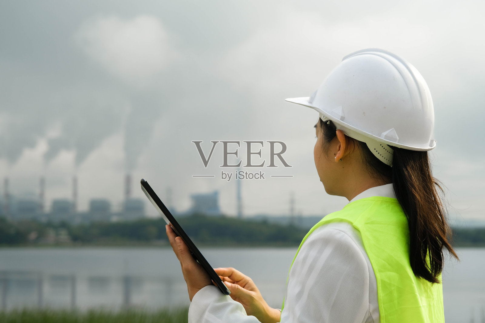 在晨雾中，身着绿色背心和头盔的女总工程师站在外面，背景是燃煤电厂站和蒸汽。在火力发电厂工作的工程师和笔记本电脑。照片摄影图片