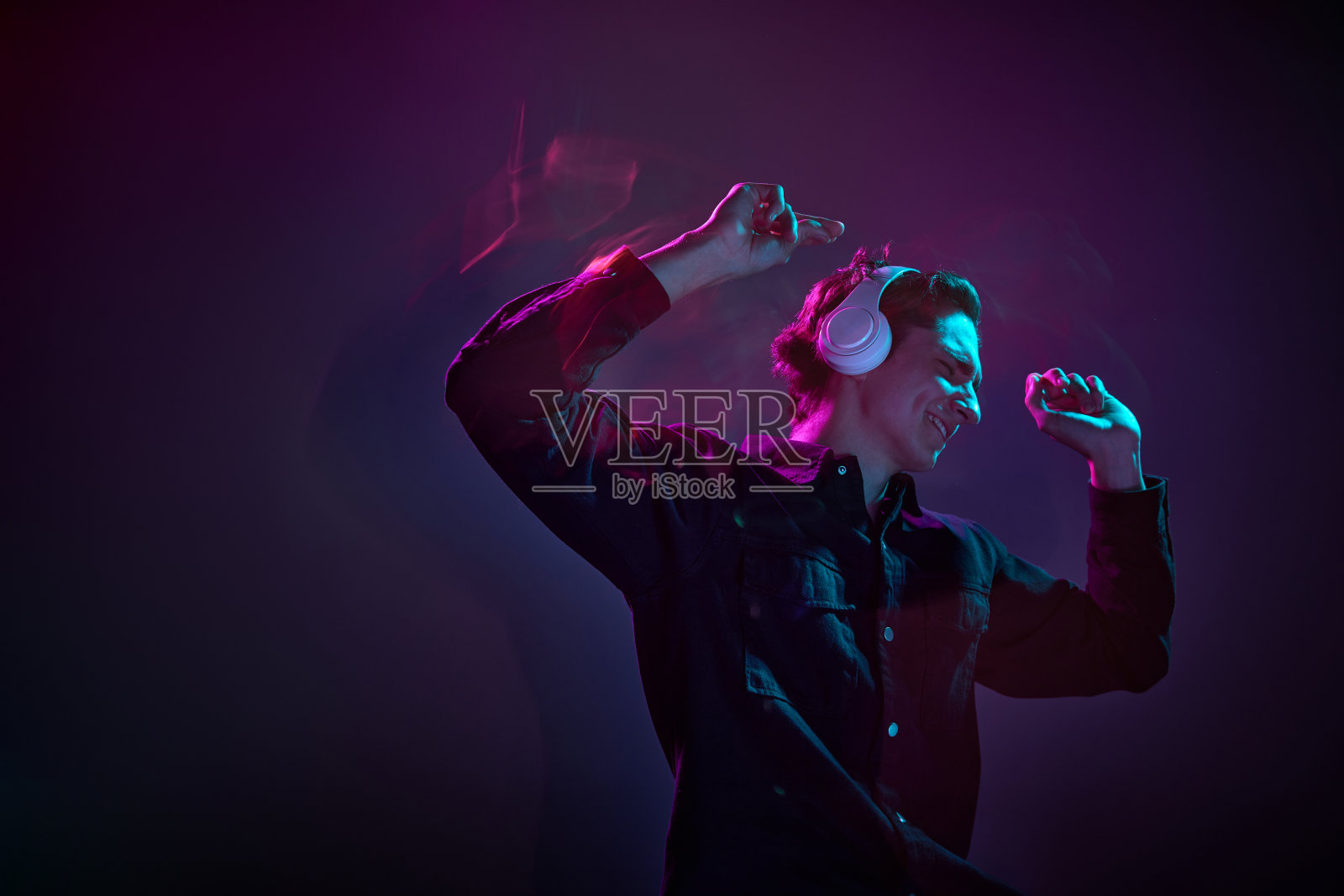 年轻男子的肖像在休闲黑色衬衫，跳舞的耳机隔离在渐变色深紫色背景在霓虹灯混合光。人类情感的概念，面部表情。照片摄影图片