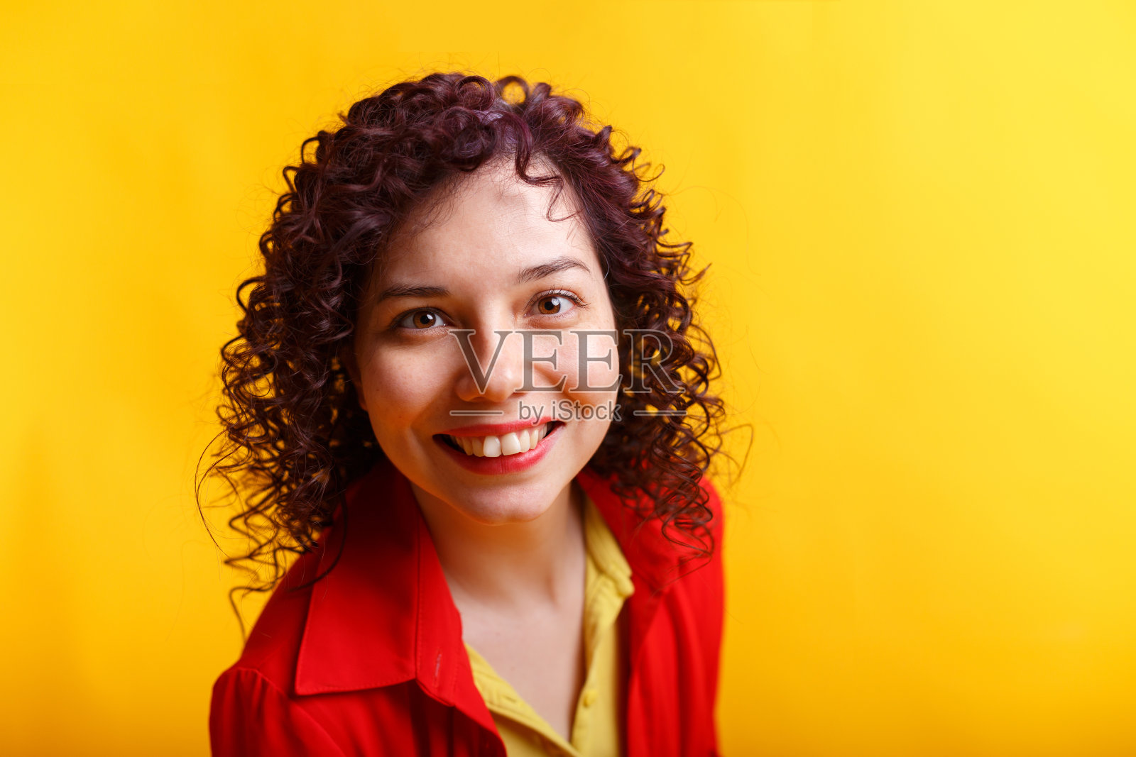 工作室拍摄的正面和微笑的女孩。穿着鲜红衬衫和黄色t恤的年轻卷发女子。黄色背景隔离。照片摄影图片