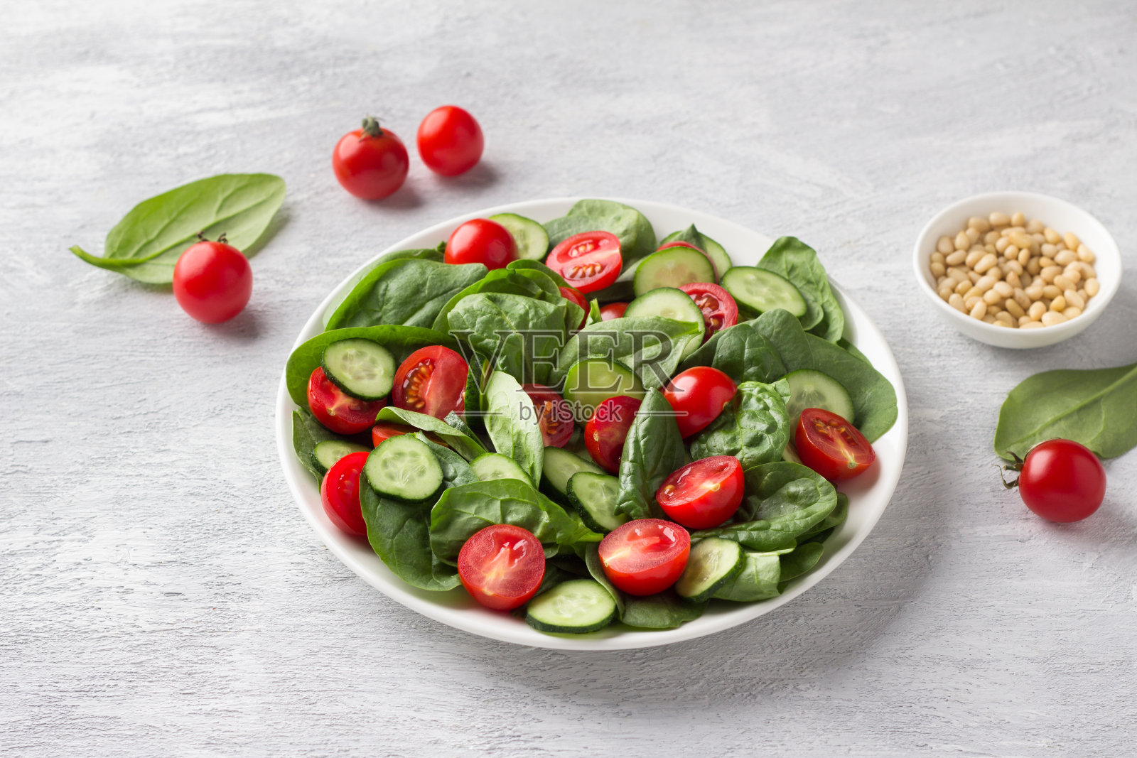 盘子里有新鲜的菠菜叶、黄瓜、圣女果和松子，背景是浅灰色的。美味健康的食物照片摄影图片