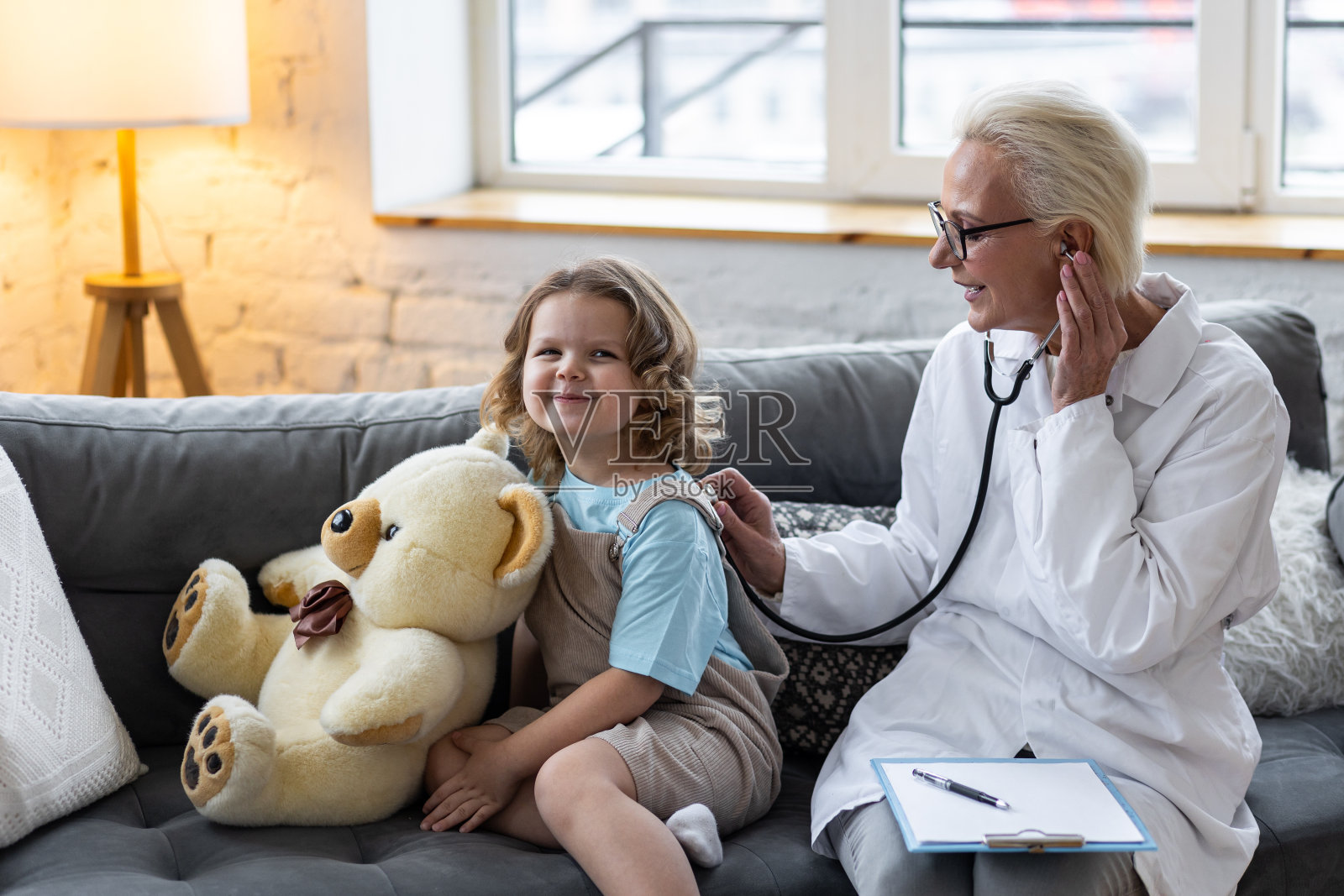 善良的女老年儿科医生到家里看望他的病人，检查小女孩，写处方。儿童健康检查的概念测量体温，心跳照片摄影图片