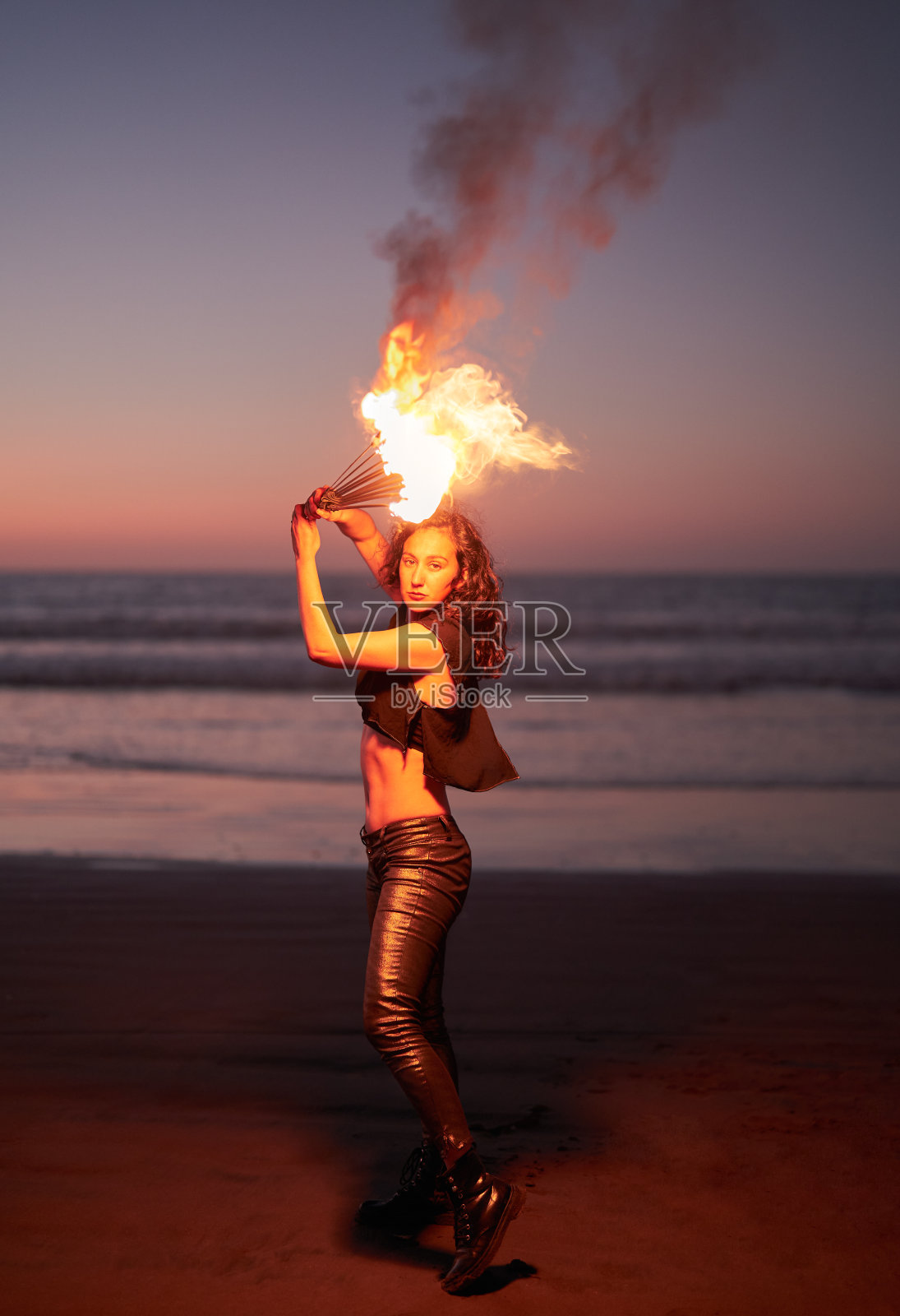 晚上，年轻的拉丁女消防演员在海滩上将火扇举过头顶照片摄影图片