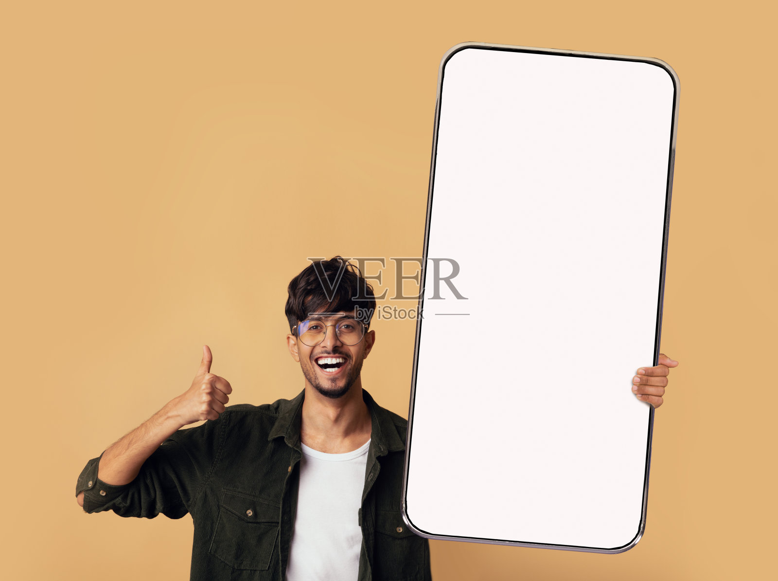 积极的年轻印度人展示大空智能手机，在米色工作室背景上竖起大拇指，模型照片摄影图片