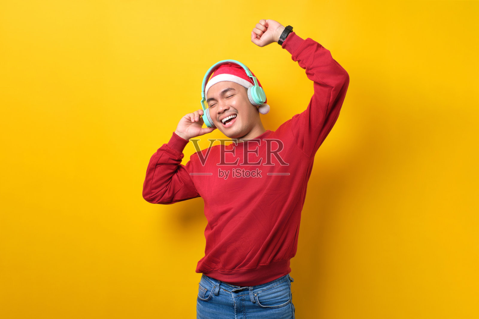 快乐的年轻亚洲人戴着圣诞帽，戴着耳机听音乐，在黄色的演播室背景上跳舞。庆祝圣诞假期和新年的概念照片摄影图片