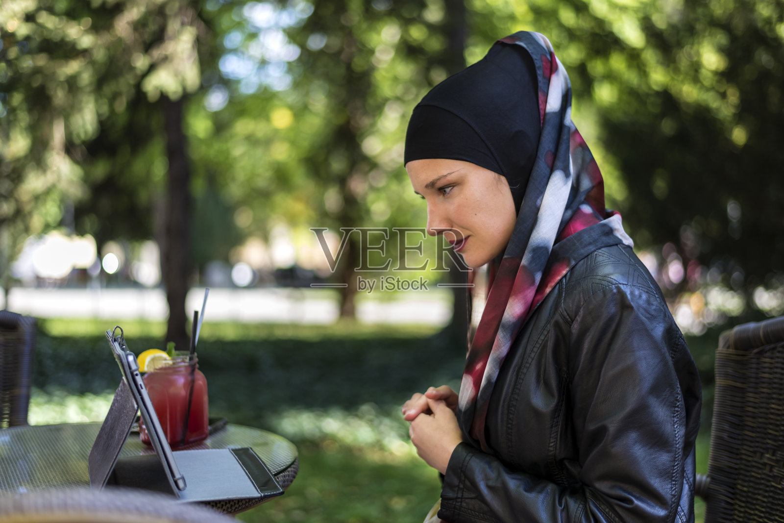 一名戴着头巾的穆斯林妇女在公园旁边的街头咖啡馆里使用平板电脑。照片摄影图片