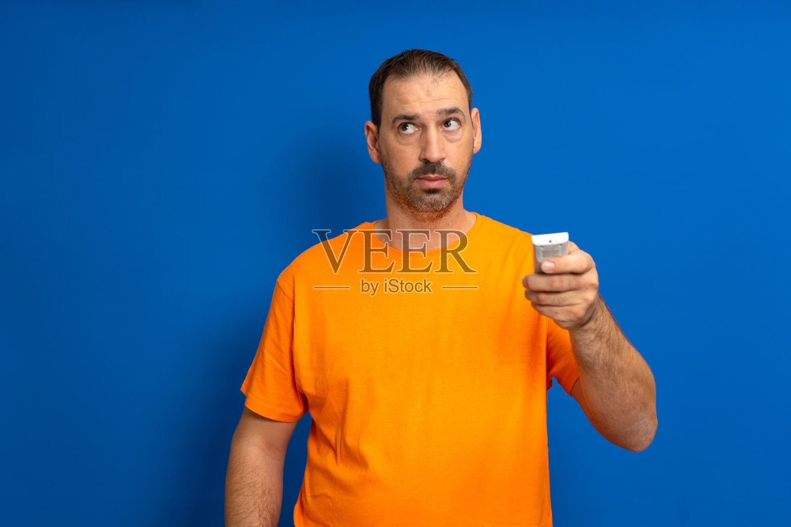 一个留着胡子、穿着橙色t恤的拉丁裔男子按下遥控器打开空调，孤立在蓝色的演播室背景上。照片摄影图片