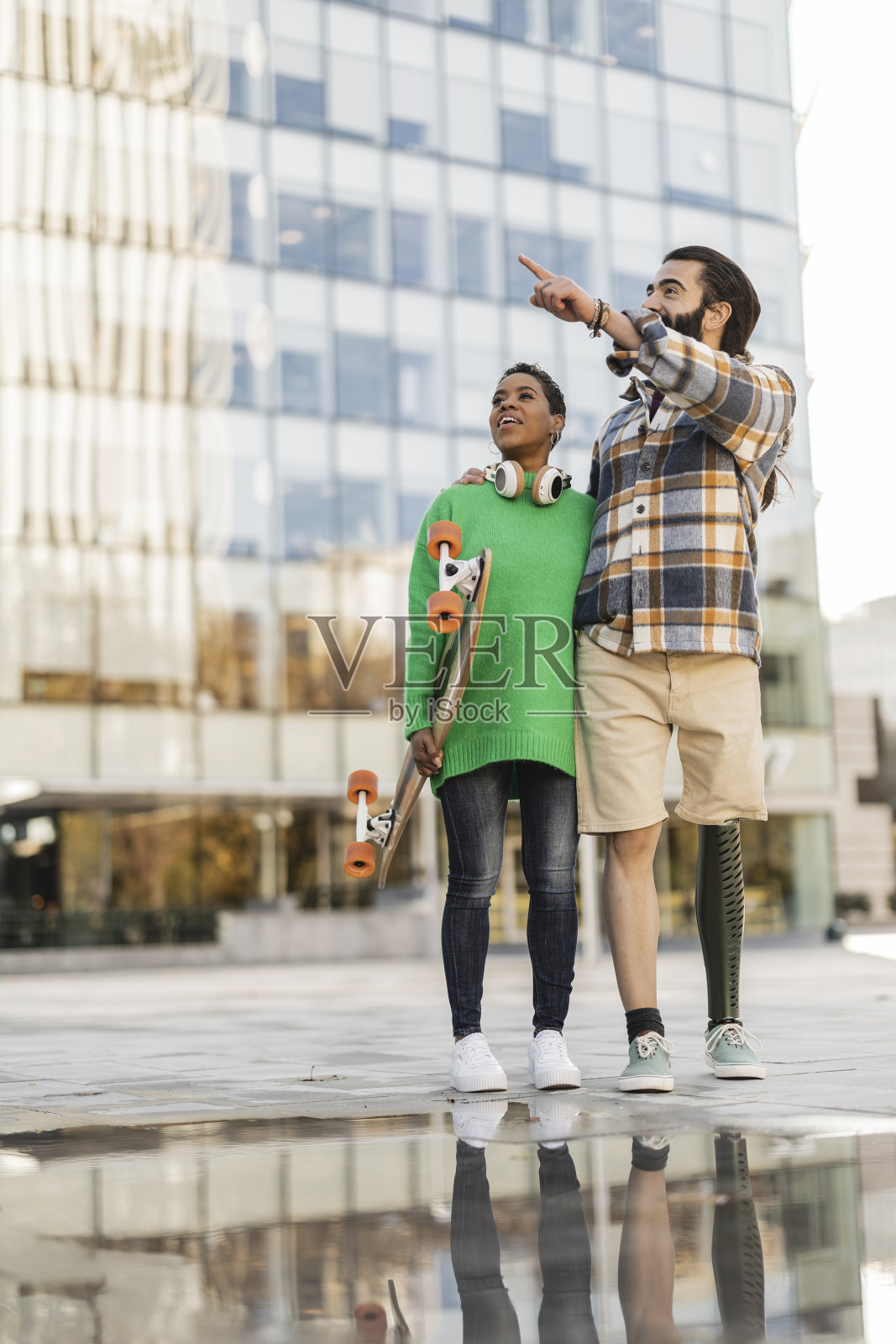 有趣的情侣带着滑板，在城市里站着指指点点，非洲裔美国女人，西班牙裔男人截肢和假肢。照片摄影图片