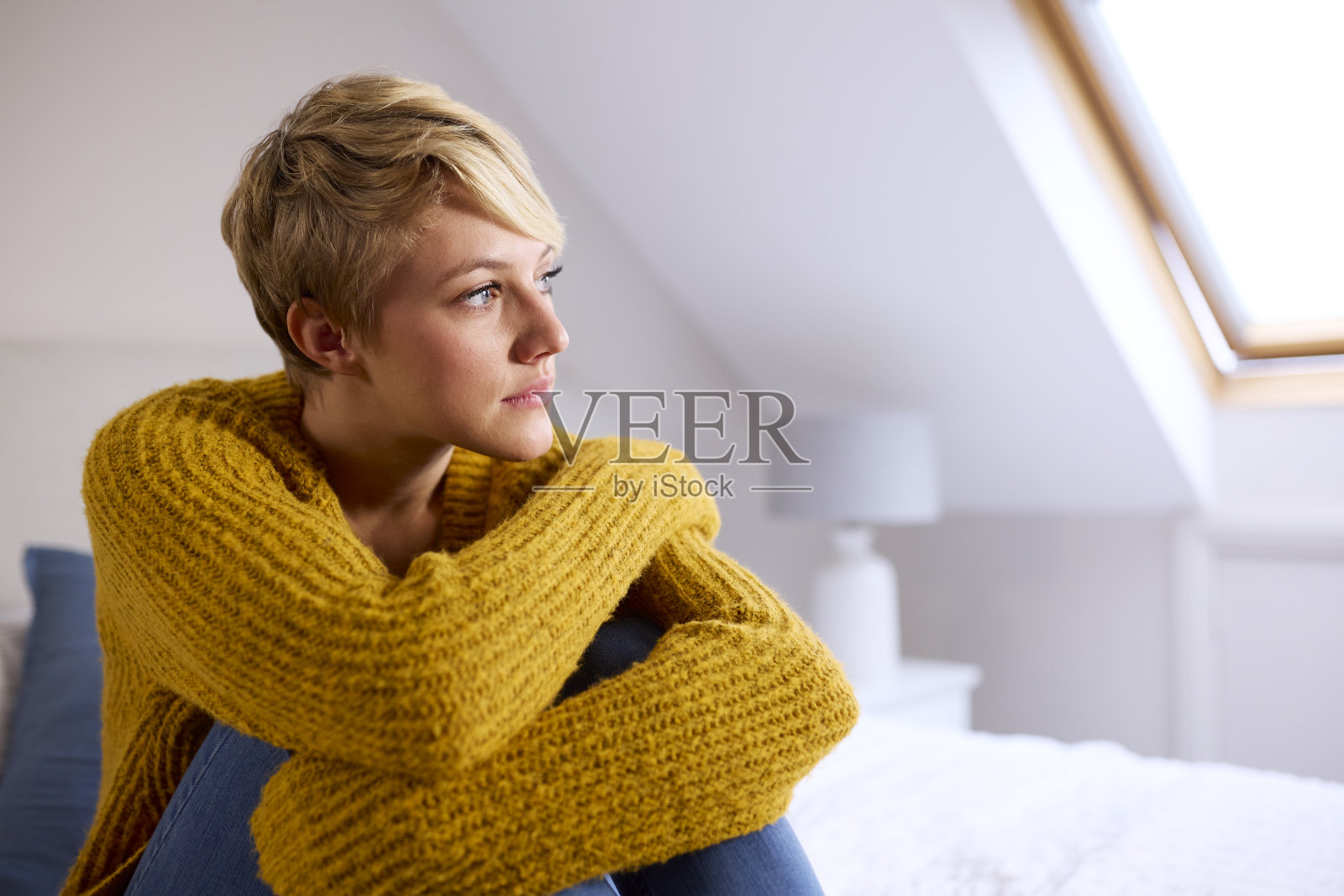 沉思的年轻女子坐在家里的床上，望着窗外照片摄影图片