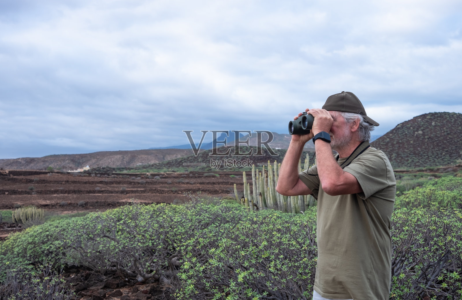 活跃的老人戴着帽子，在大自然中享受徒步旅行，站在双筒望远镜看着山景，退休老人享受假期和自由照片摄影图片