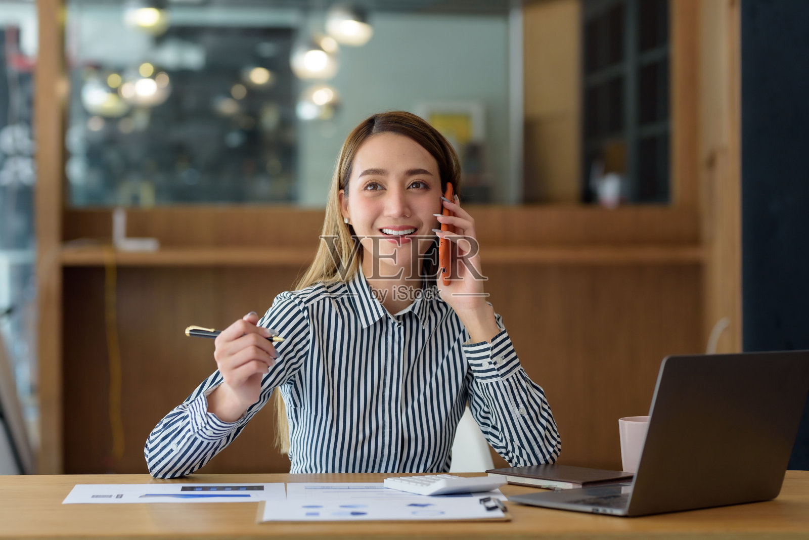 微笑的年轻亚洲女商人坐在办公室的桌子上用笔记本电脑打电话。照片摄影图片