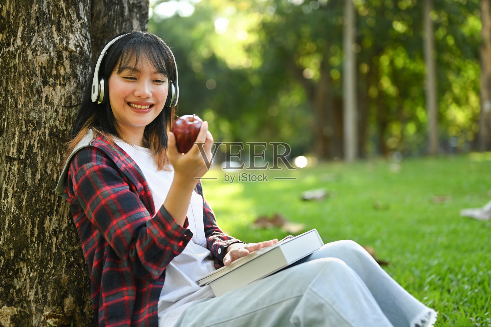 漂亮的年轻亚洲女子在阳光明媚的美丽花园背景下的绿草地上的大树下吃苹果。照片摄影图片