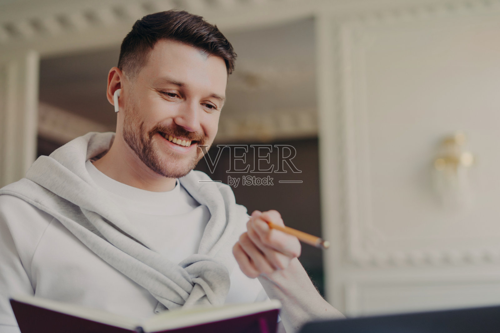 穿着套头衫和无线耳机的微笑男子自由职业者，手里拿着铅笔和书，在笔记本电脑上进行视频通话照片摄影图片