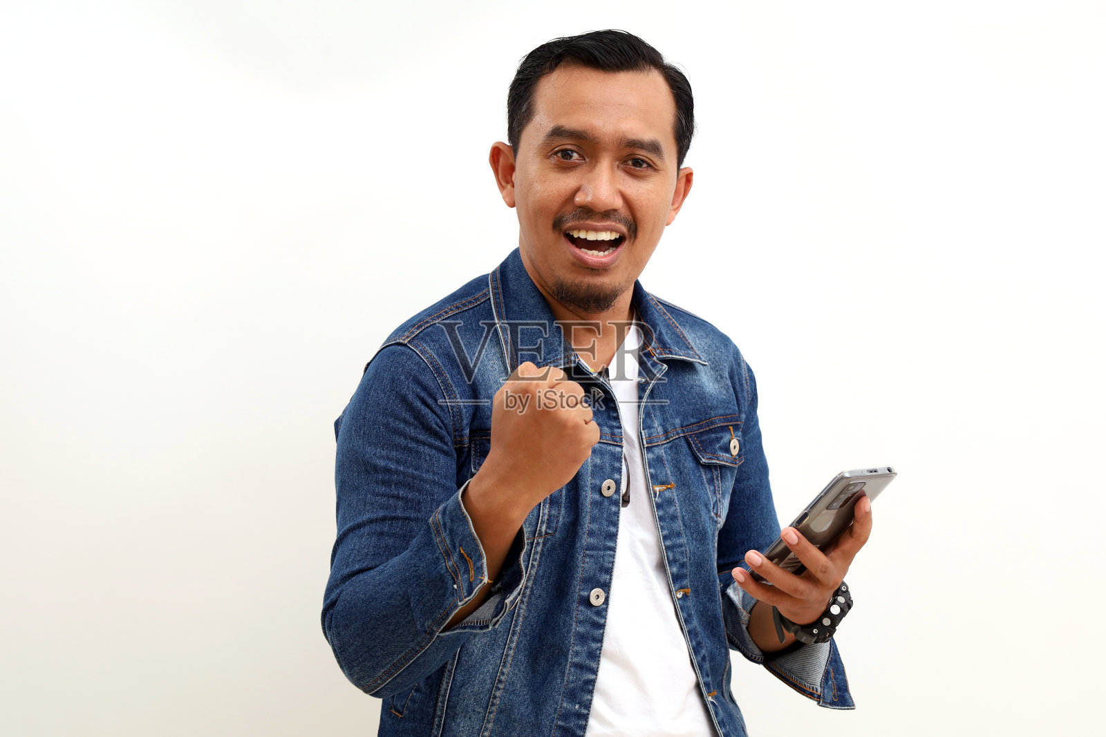 快乐的亚洲男人站着，拿着手机，拳头他的手。白色背景隔离照片摄影图片