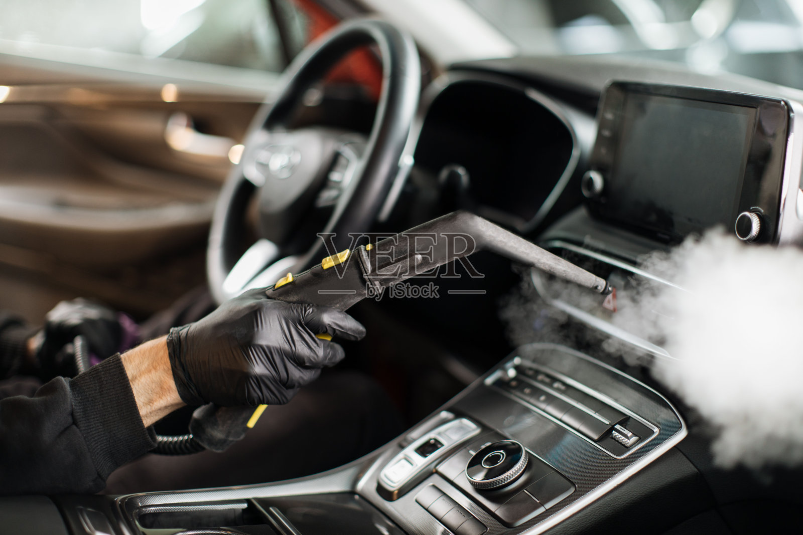 戴着黑色橡胶手套的男子用热蒸汽清洗机清洗汽车内部。照片摄影图片