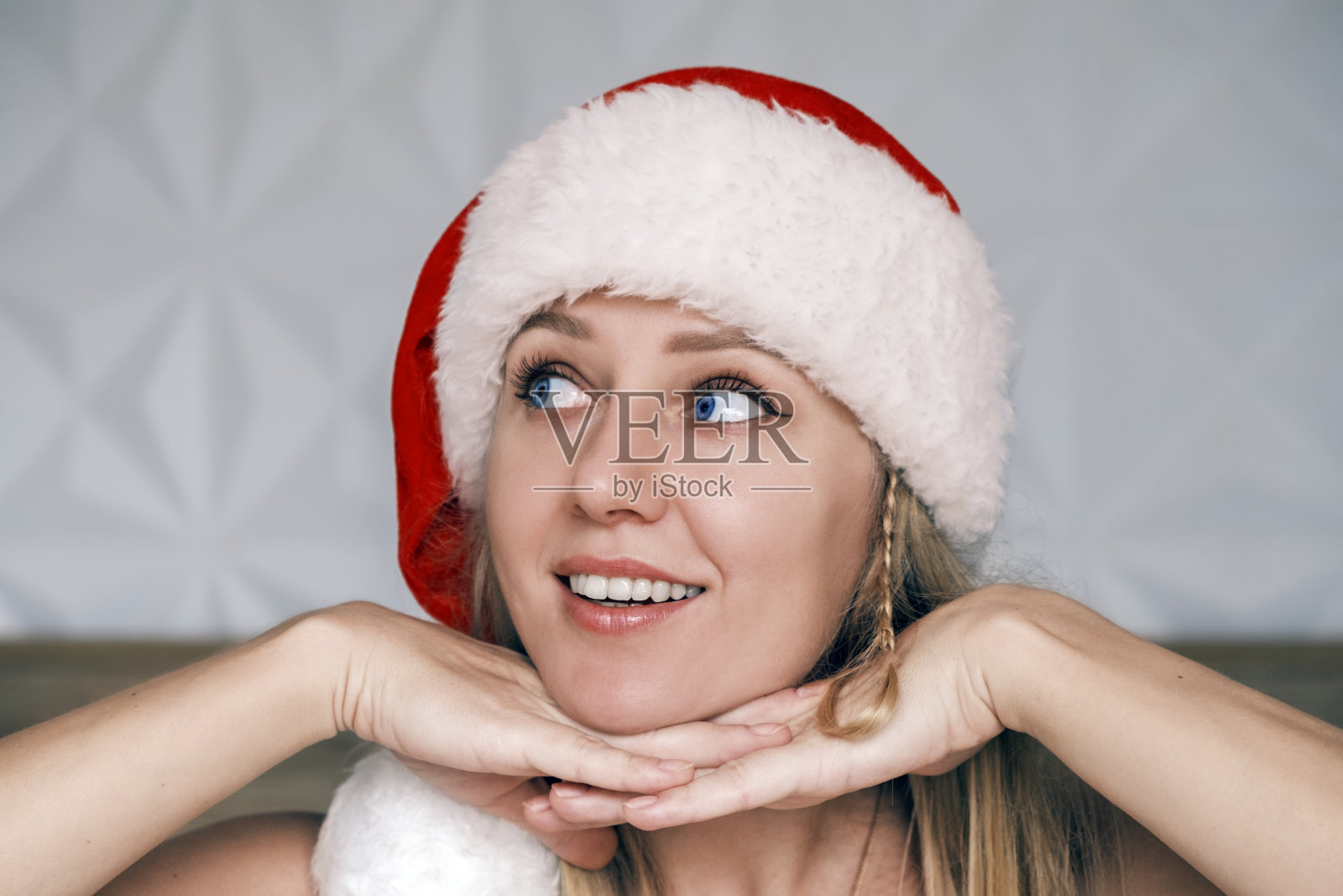 戴着红帽子的圣诞女孩微笑着，双手托着下巴向上看。戴着圣诞老人帽子的漂亮的年轻蓝眼睛金发女人的肖像。圣诞节的肖像。圣诞假期，假期照片摄影图片