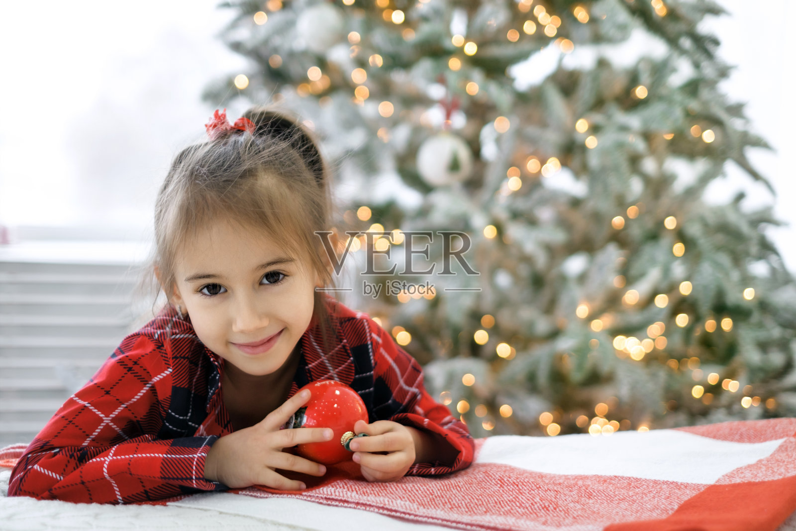 一个女孩的肖像在格纹睡衣与一个圣诞球在手中。孩子坐在圣诞树旁边的床上照片摄影图片