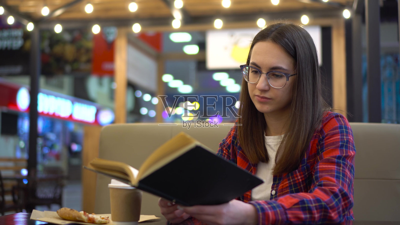 一位年轻女子正在一家舒适的咖啡馆里看书。一个女人坐在餐桌边吃着披萨。穿红衬衫戴眼镜的女孩。照片摄影图片