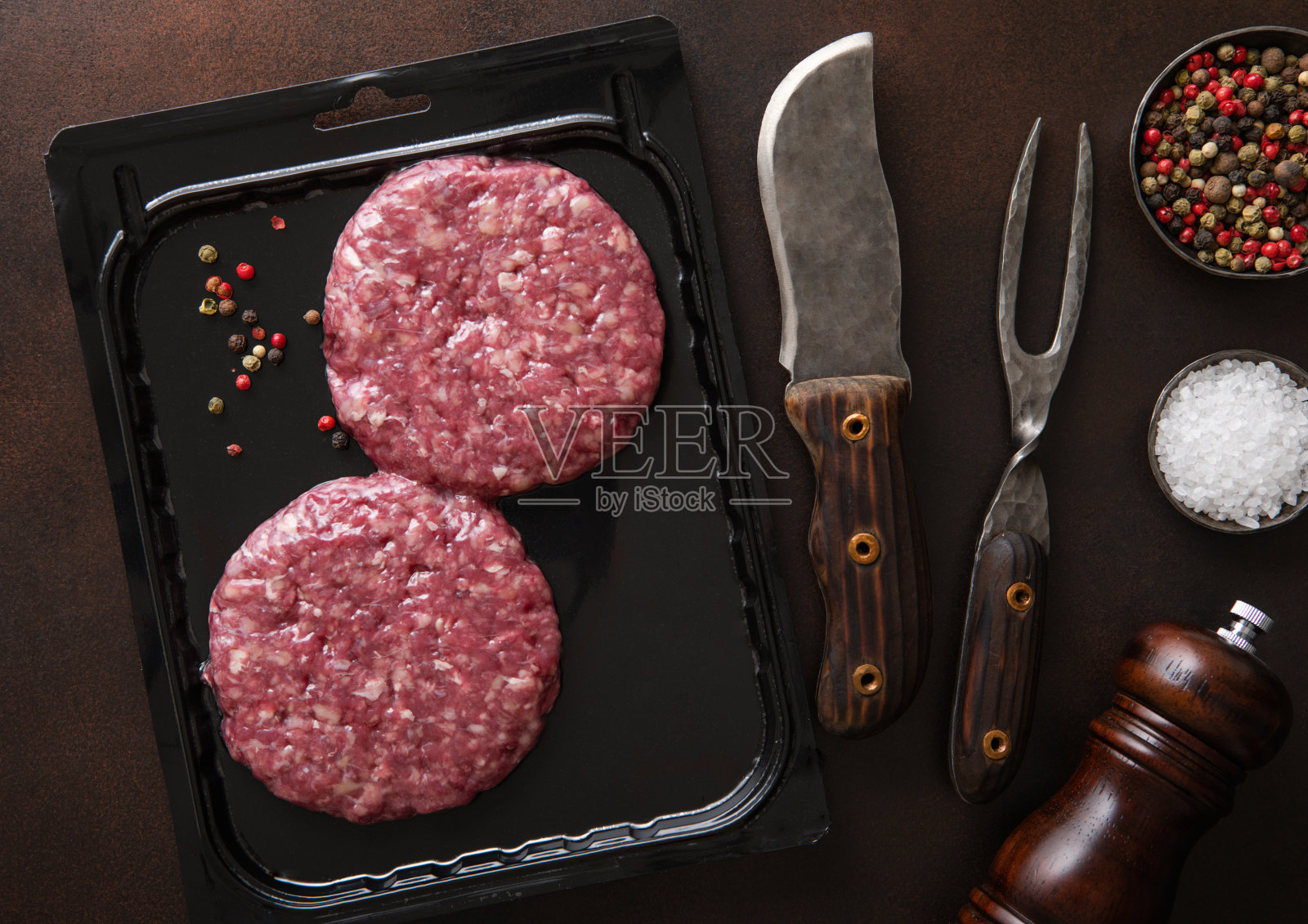 生牛肉汉堡密封在真空盘与烧烤叉和刀在黑暗的厨房背景与胡椒研磨机。照片摄影图片