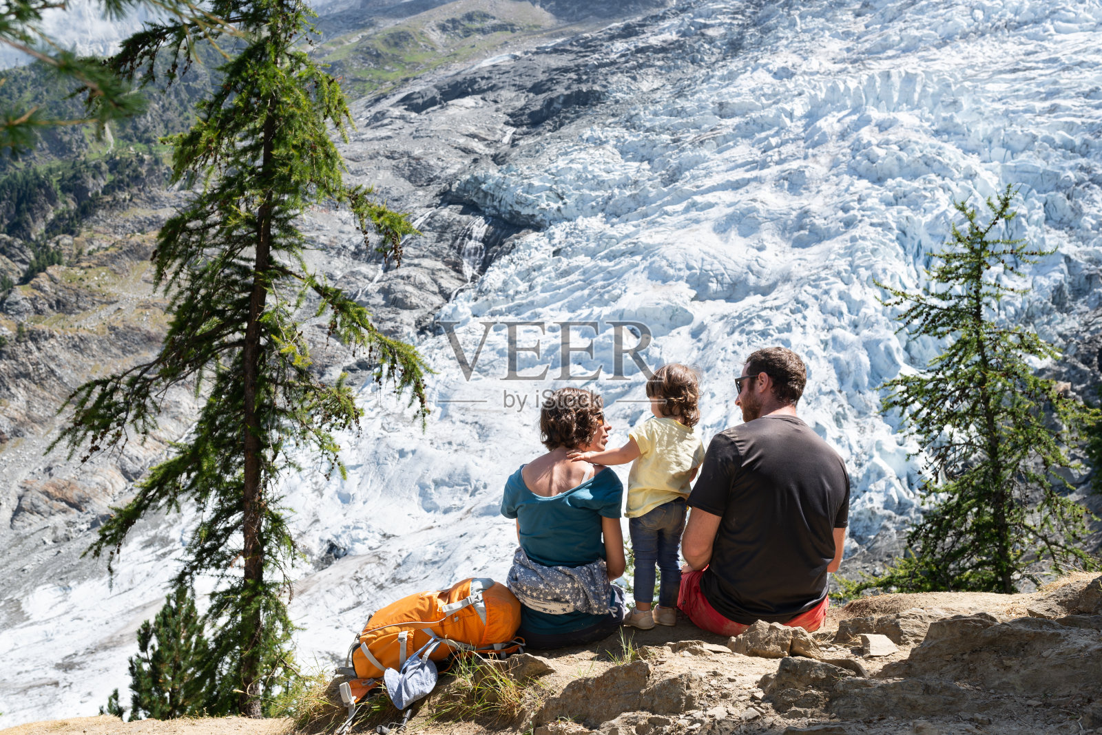 一对夫妇带着蹒跚学步的孩子坐在岩石上，欣赏法国阿尔卑斯山的博松冰川。照片摄影图片
