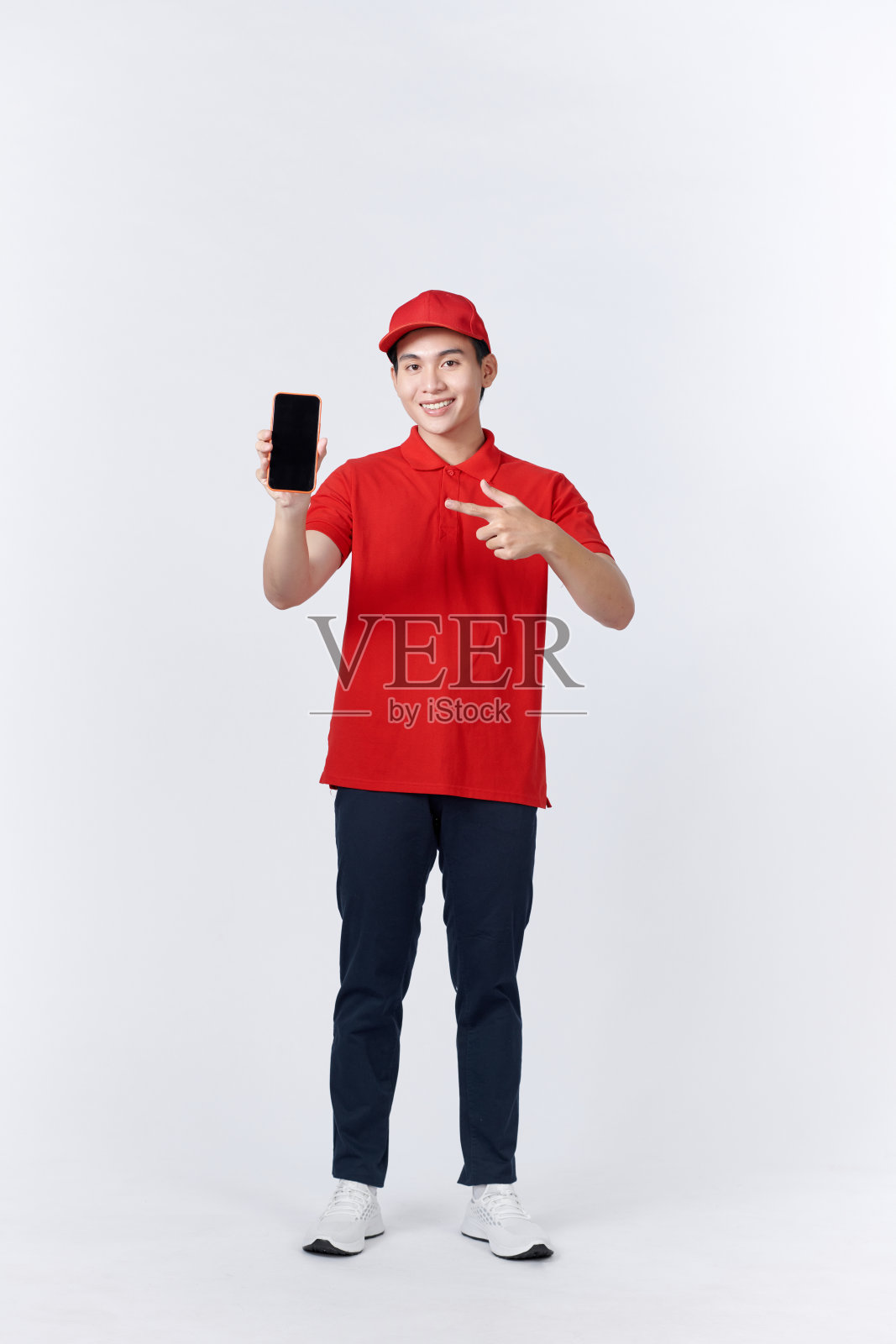 亚洲快递员戴着帽子，拿着智能手机，白色背景，非常高兴地用手和手指指着一边照片摄影图片