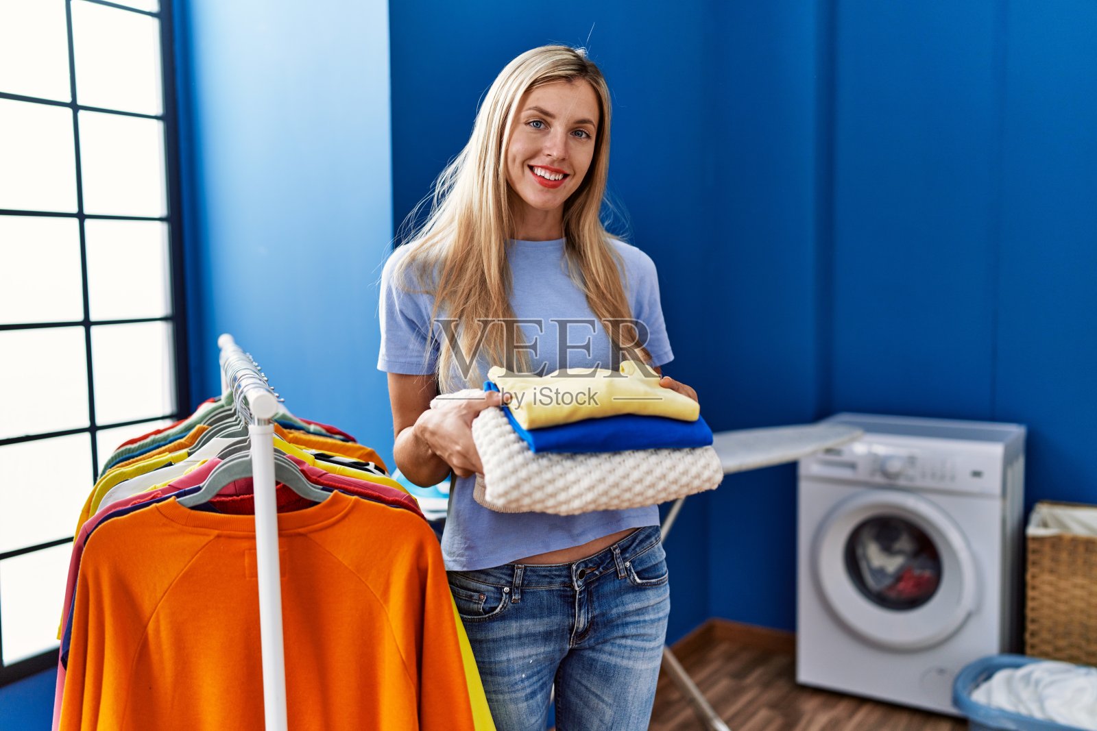 年轻的金发女郎微笑着自信地拿着叠好的衣服在洗衣房照片摄影图片