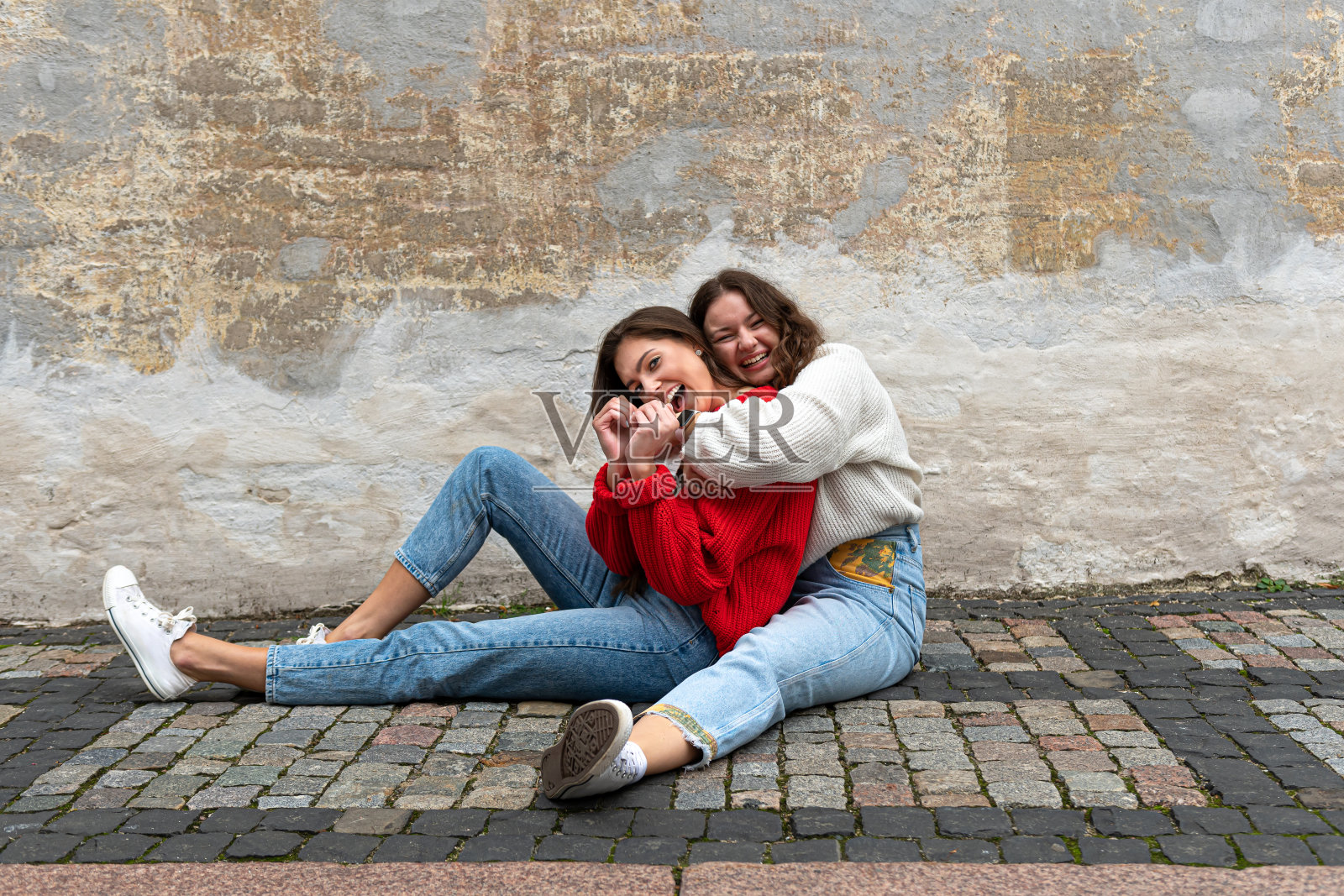 两名年轻女子抱在一起，坐在一堵旧墙旁的人行道上照片摄影图片