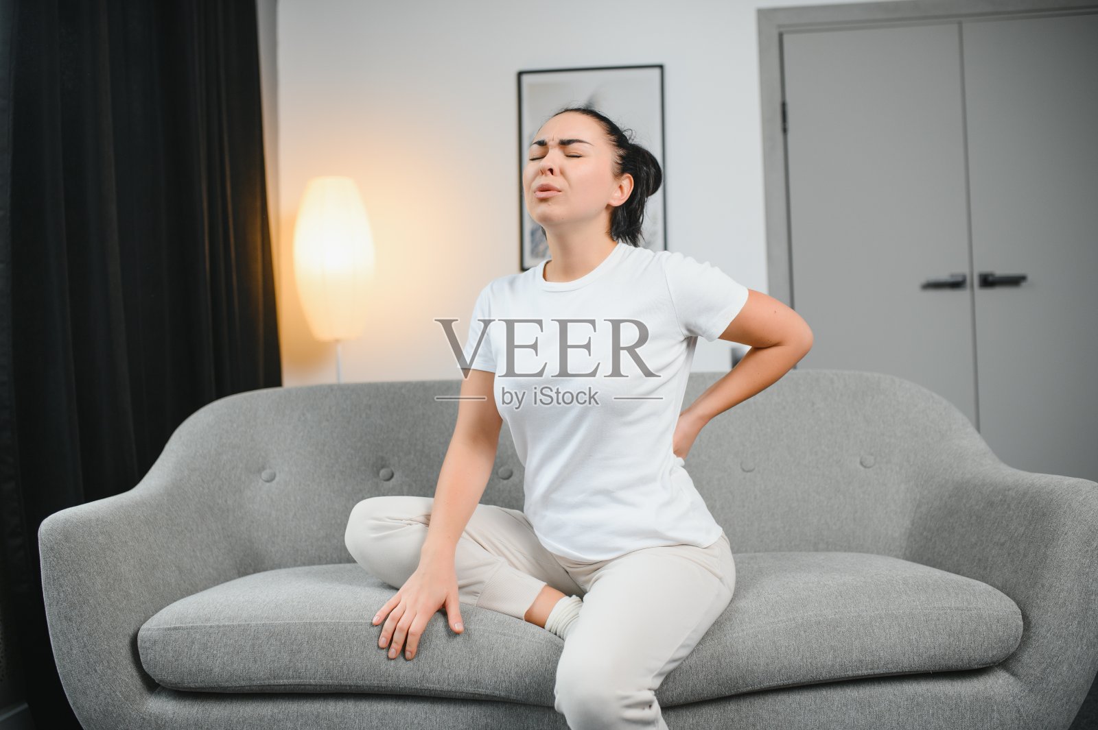 漂亮的女孩坐在被褥上，闭着眼睛，忍受着背痛。照片摄影图片