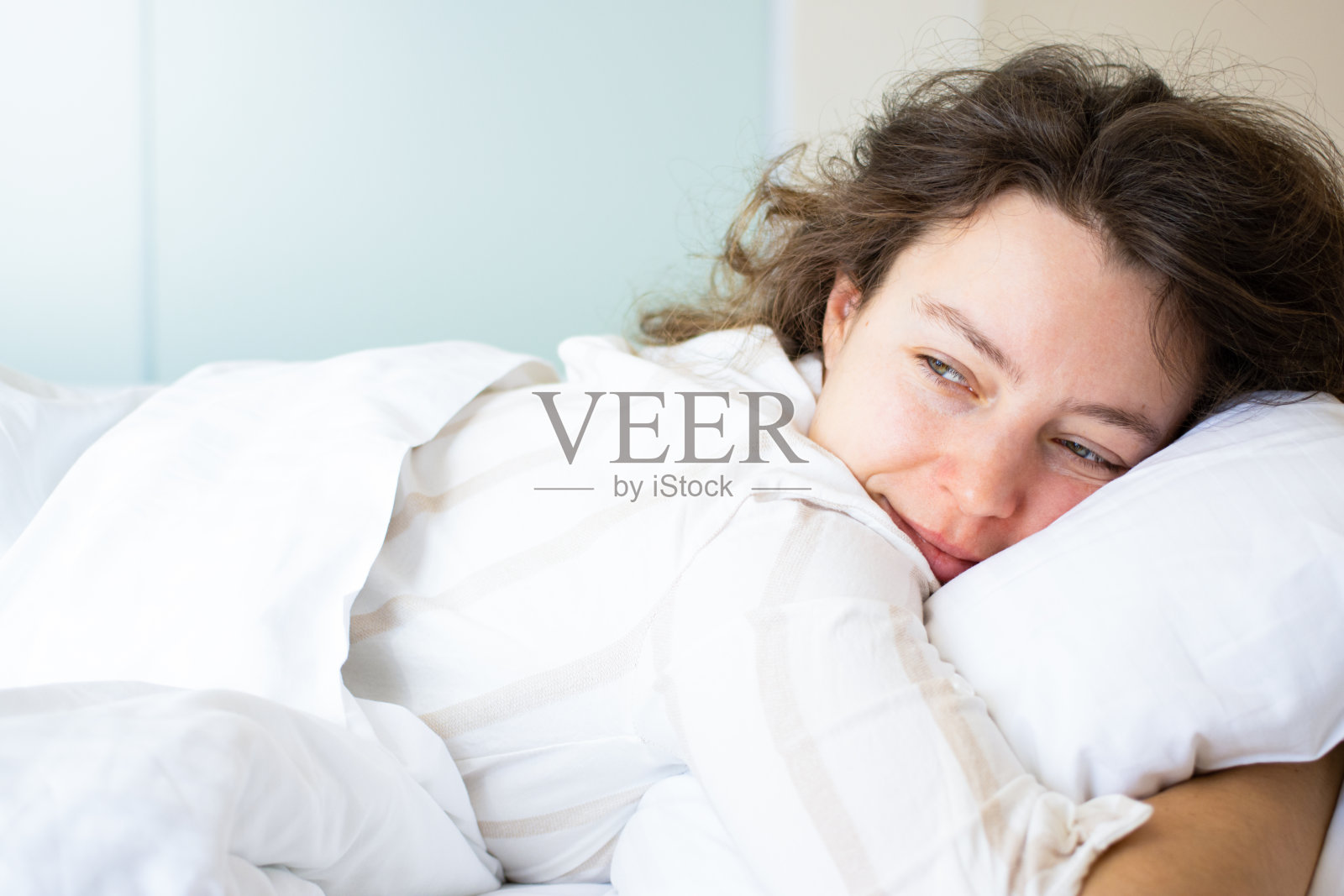 幸福的黑发女人躺在床上拥抱柔软的白色枕头。微笑遥望，享受卧室里新鲜柔软的床上用品和矫形床垫。放松，休息，慵懒的周末早晨。照片摄影图片