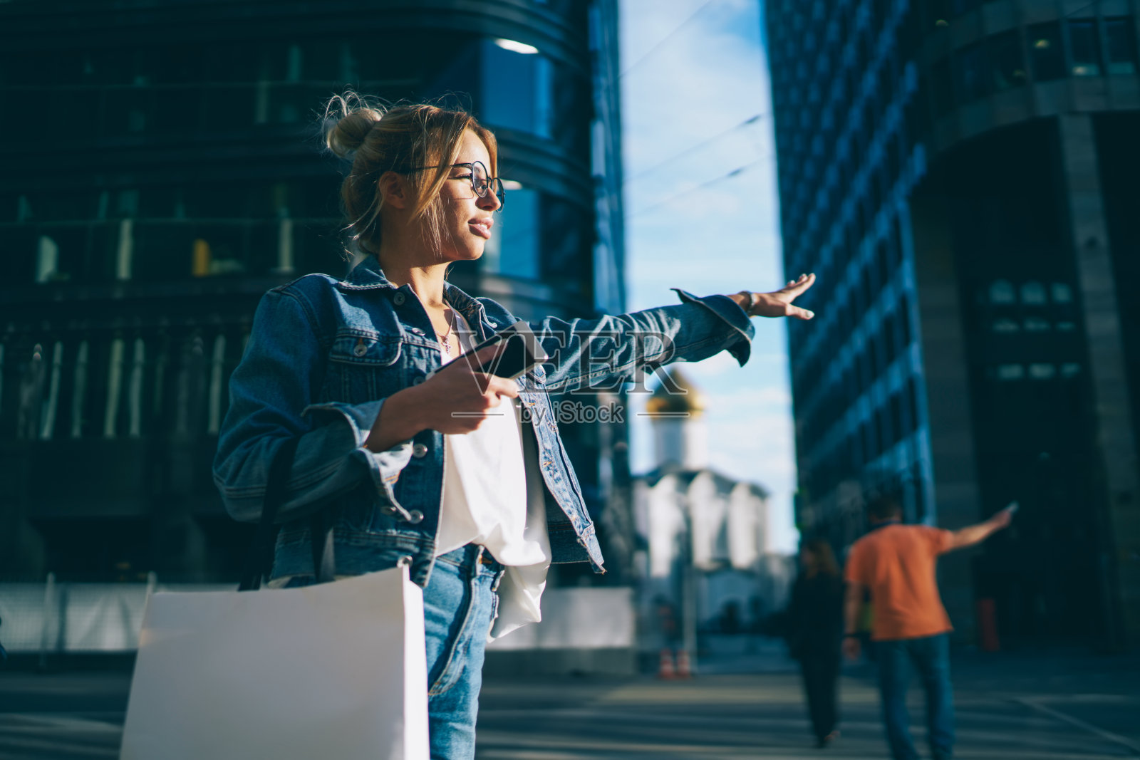 时髦时髦的女孩在光学眼镜与时髦的衣服在包装与模拟区域停止出租车站在户外。迷人的女性穿着牛仔装在市中心追赶汽车照片摄影图片